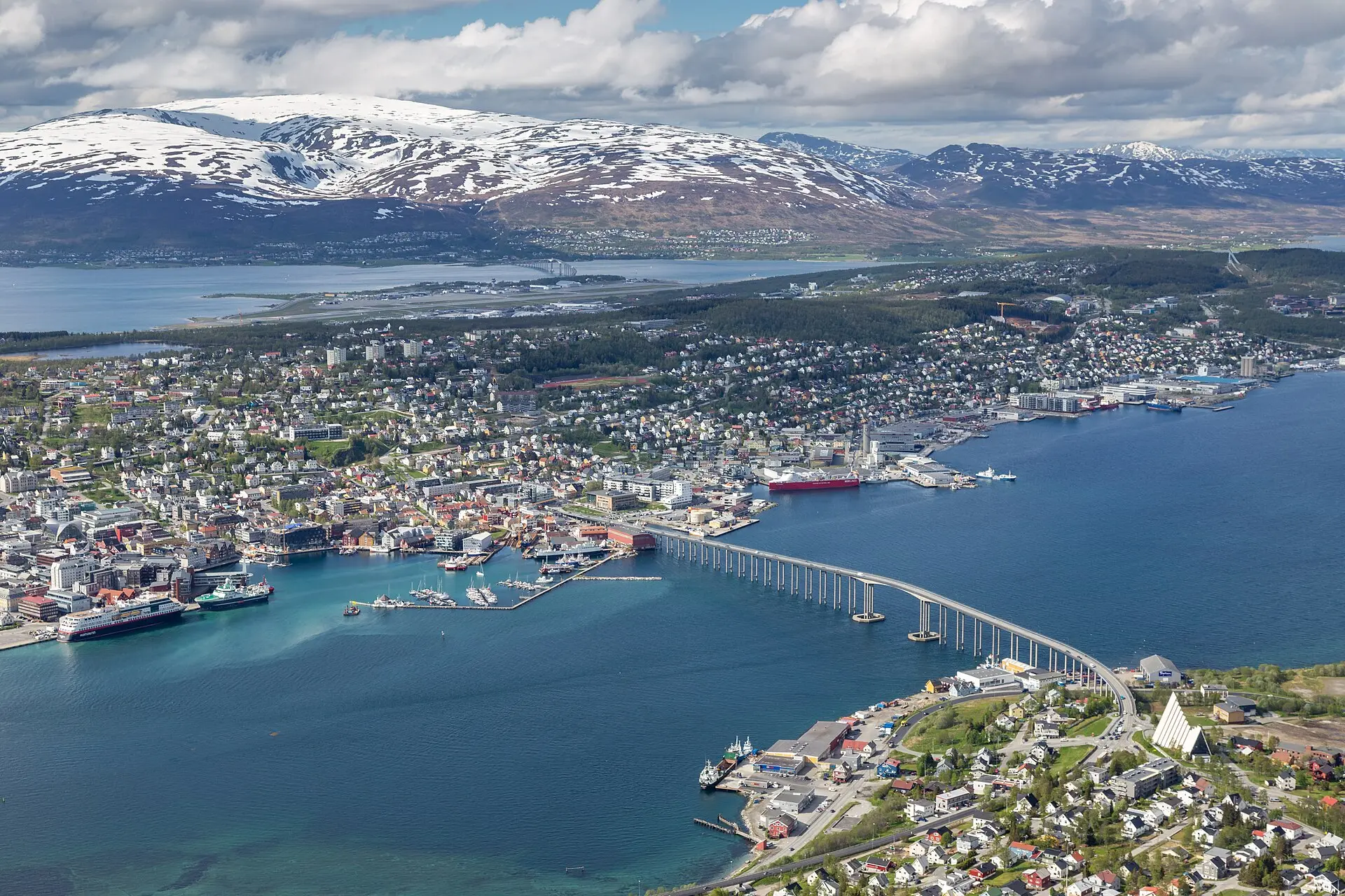 Tromsø von oben mit der Brücke Tromsøbrua, der weißen Eismeerkathedrale, Schiffen an den Kais und Bergen mit Schnee