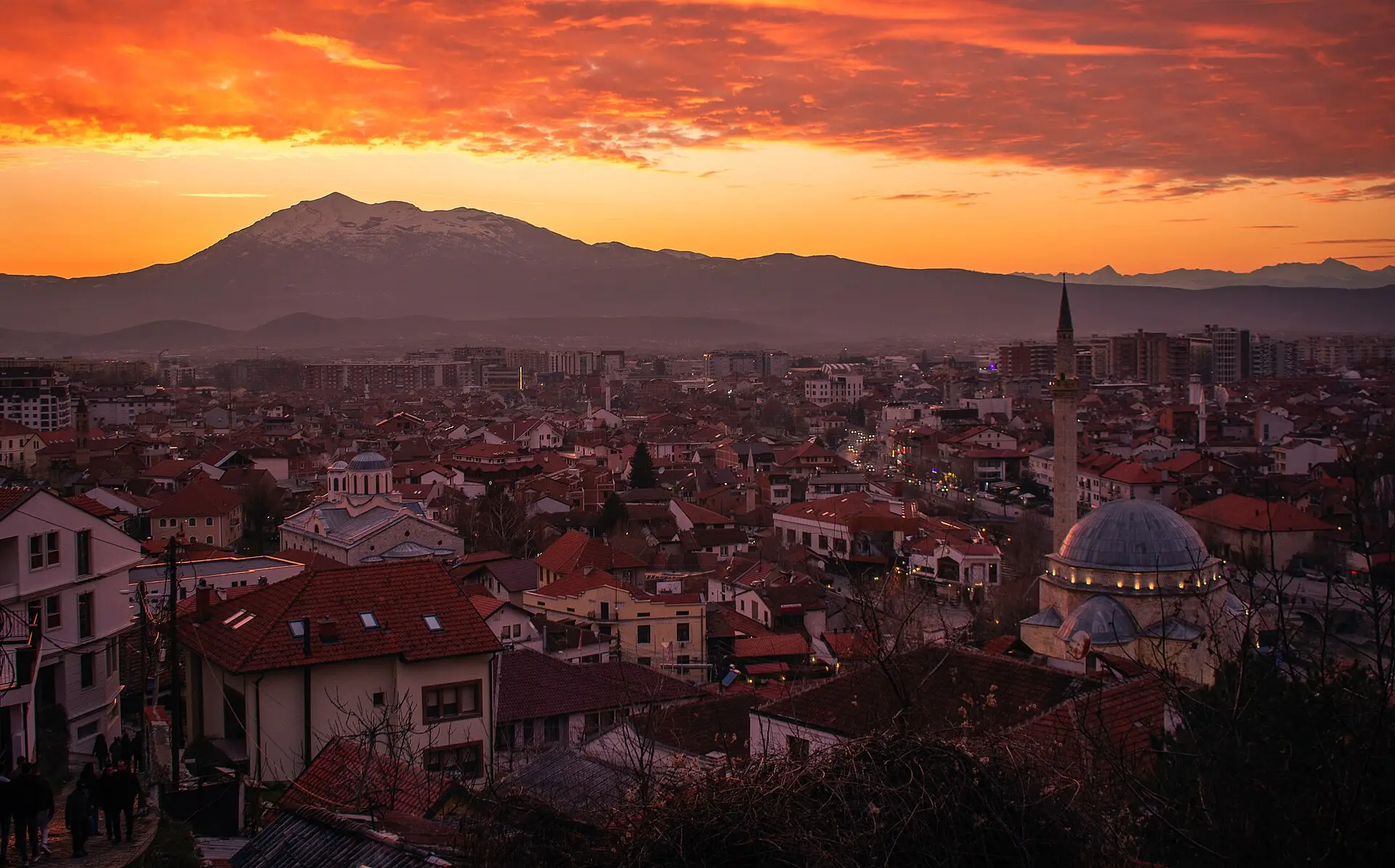 Ziegeldächer von Prizren im Abendrot, rechts die Sinan-Pascha-Moschee mit Kuppel und schlankem Minarett, dahinter ein verschneiter Berg