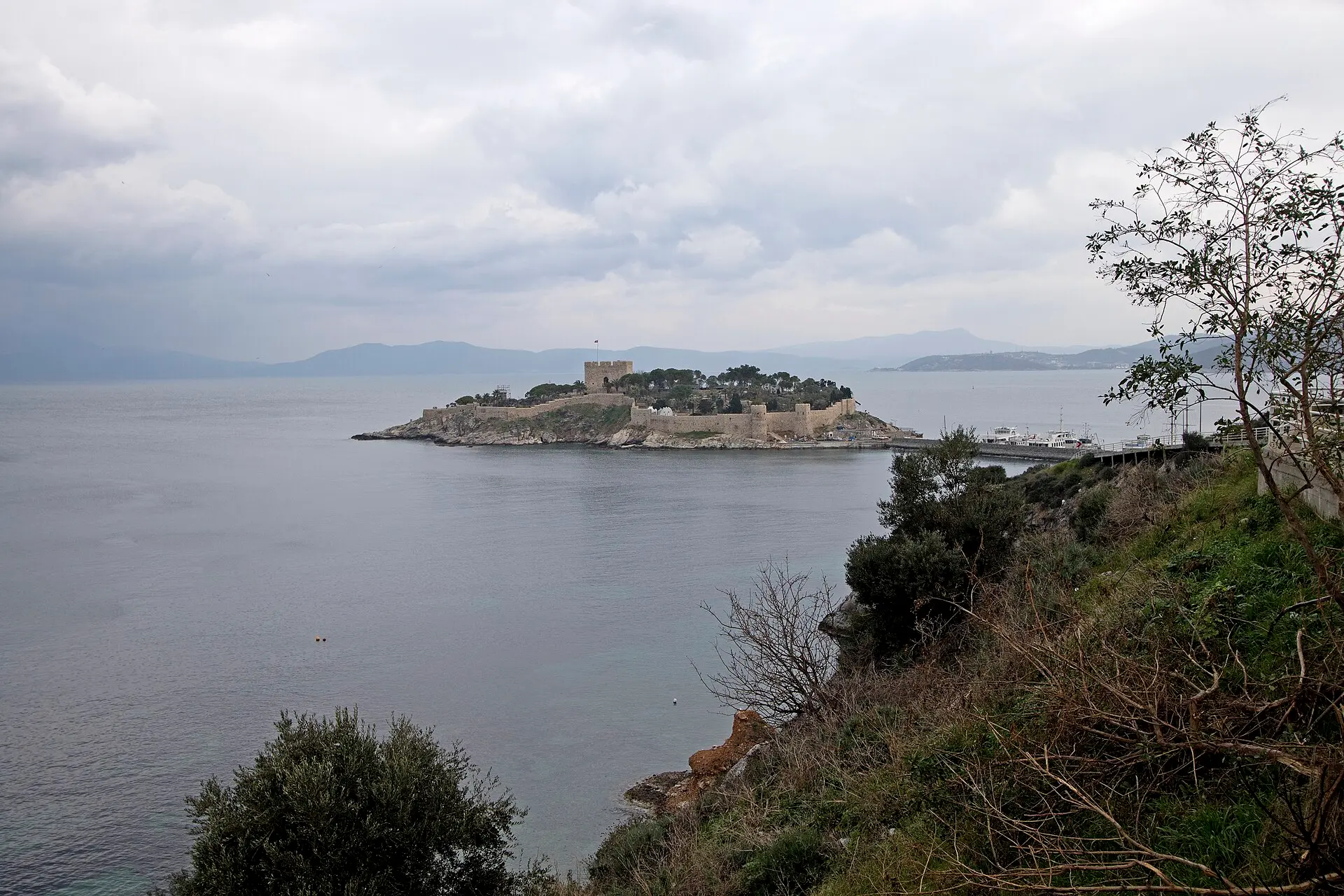 Taubeninsel vor Kuşadası mit Burgmauern und Turm, graues Meer unter Wolken, rechts ein Anleger mit Booten