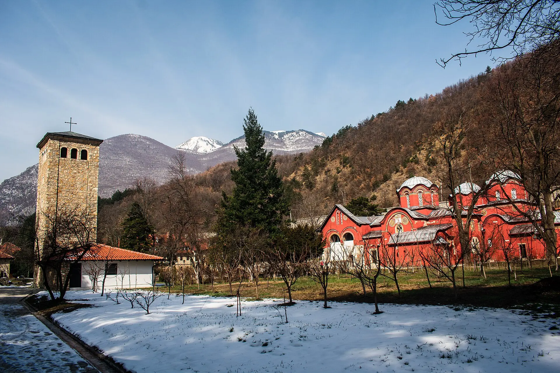 Rot verputzte Kuppelkirchen des Patriarchatsklosters Peć zwischen kahlen Bäumen, davor Schnee, dahinter verschneite Gipfel