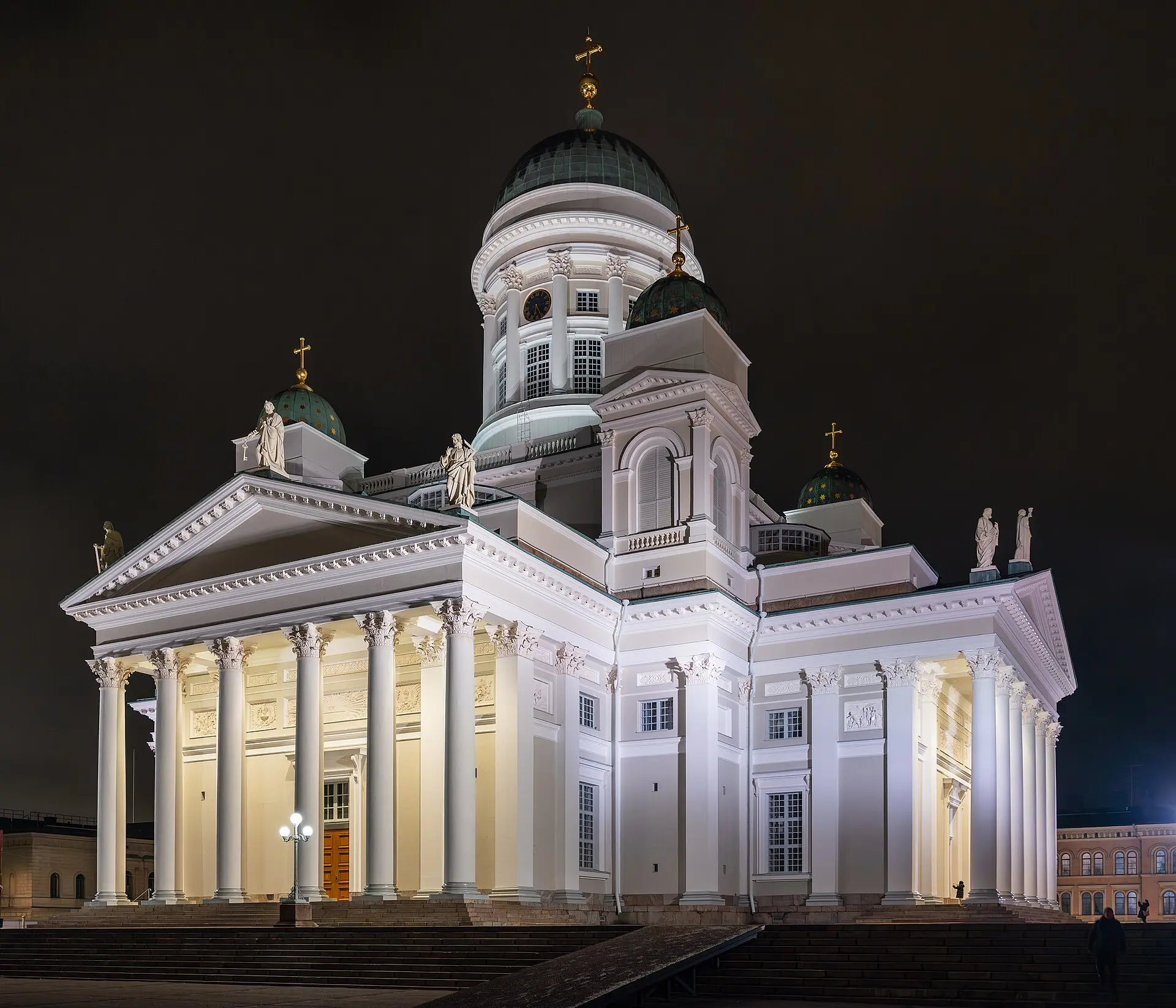 Der Dom von Helsinki bei Nacht: weiße Säulenfassade, grüne Kuppeln und Apostelfiguren hell angestrahlt, davor breite Stufen