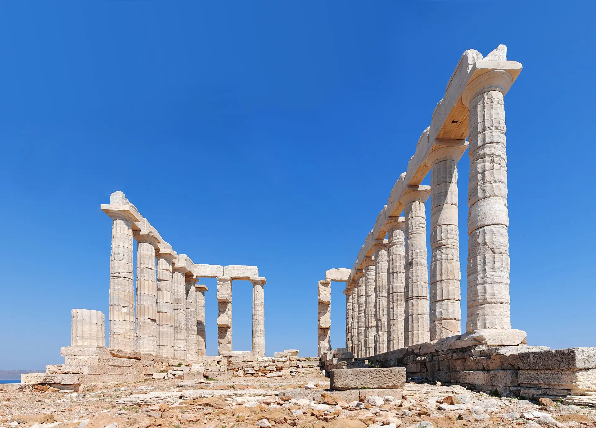 Säulenreihen des Poseidon-Tempels am Kap Sounion vor tiefblauem Himmel, Felsboden im Vordergrund, links ein Streifen Meer