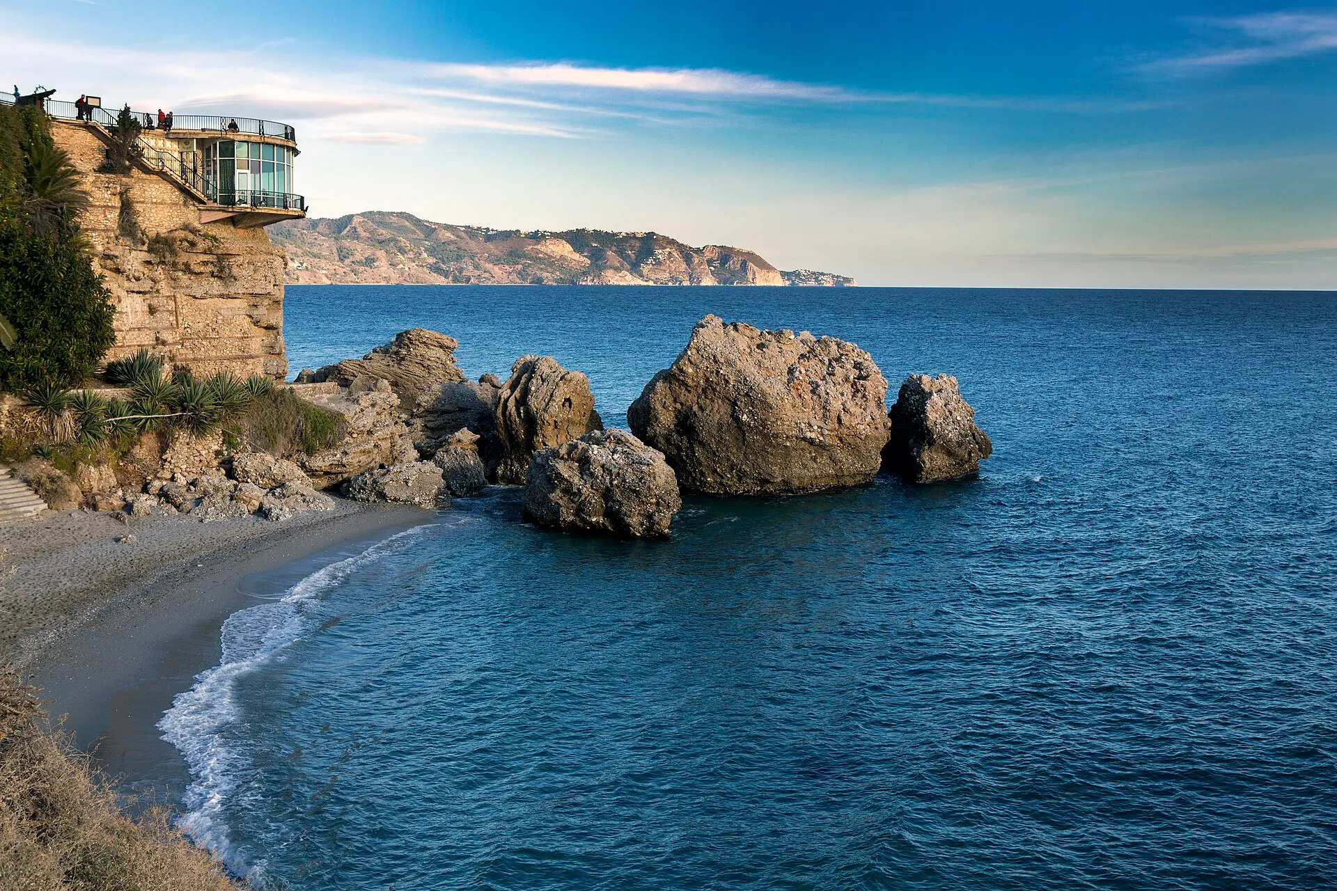 Aussichtsplattform Balcón de Europa in Nerja auf Felsen, darunter ein kleiner Sandstrand und Felsblöcke im Meer, dahinter Berge