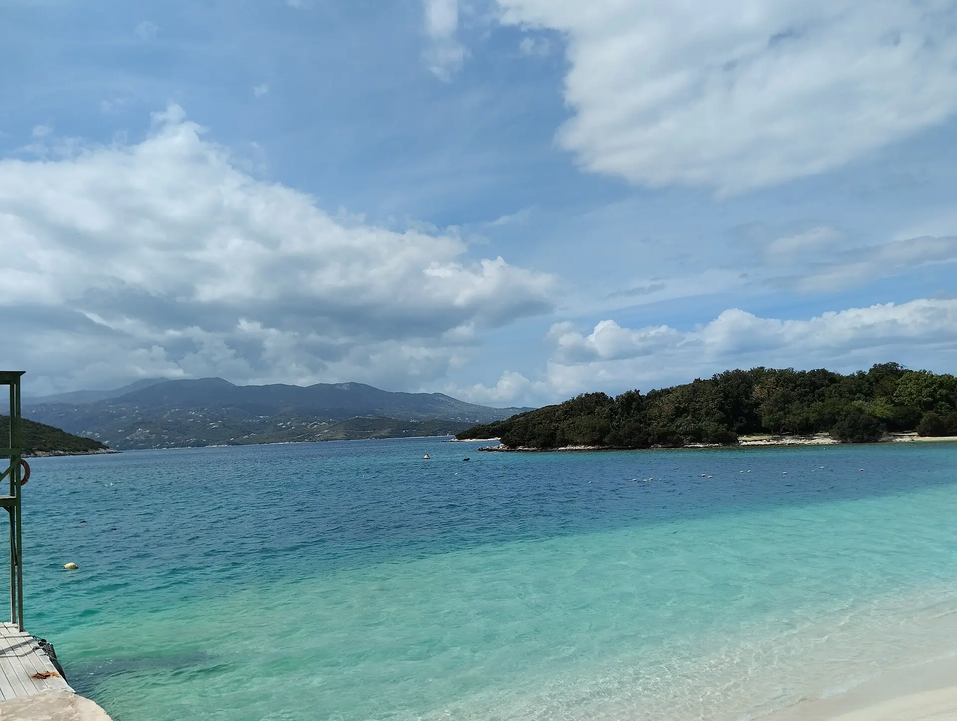 Türkises, flaches Wasser mit hellem Sandufer bei Ksamil, rechts eine bewaldete kleine Insel, hinten die Berge von Korfu