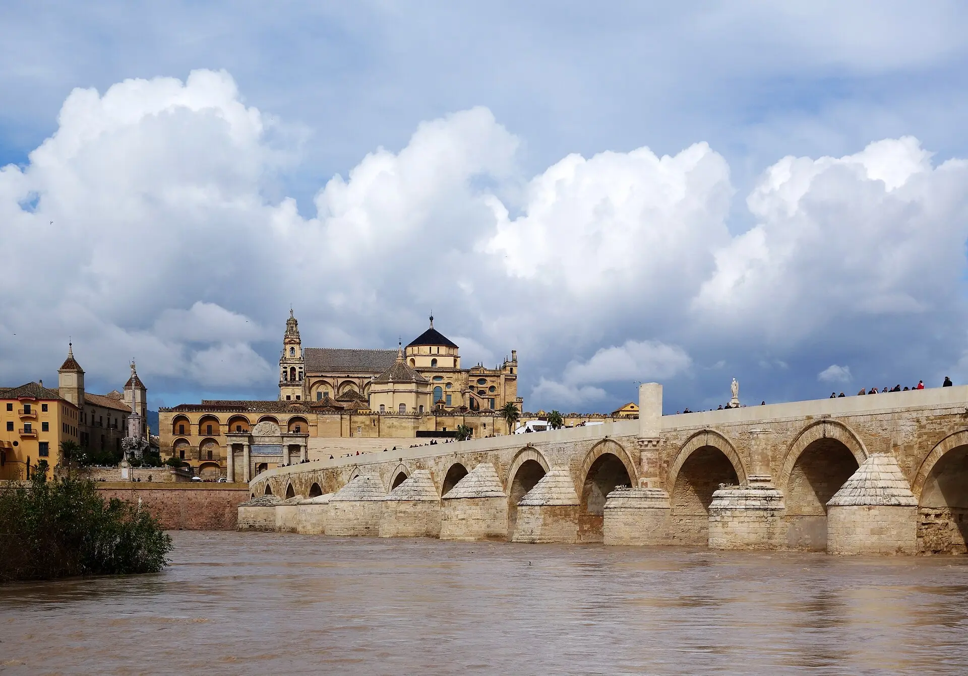 Römische Brücke mit vielen Bögen über den braunen Guadalquivir in Córdoba, dahinter die Mezquita-Kathedrale mit Glockenturm