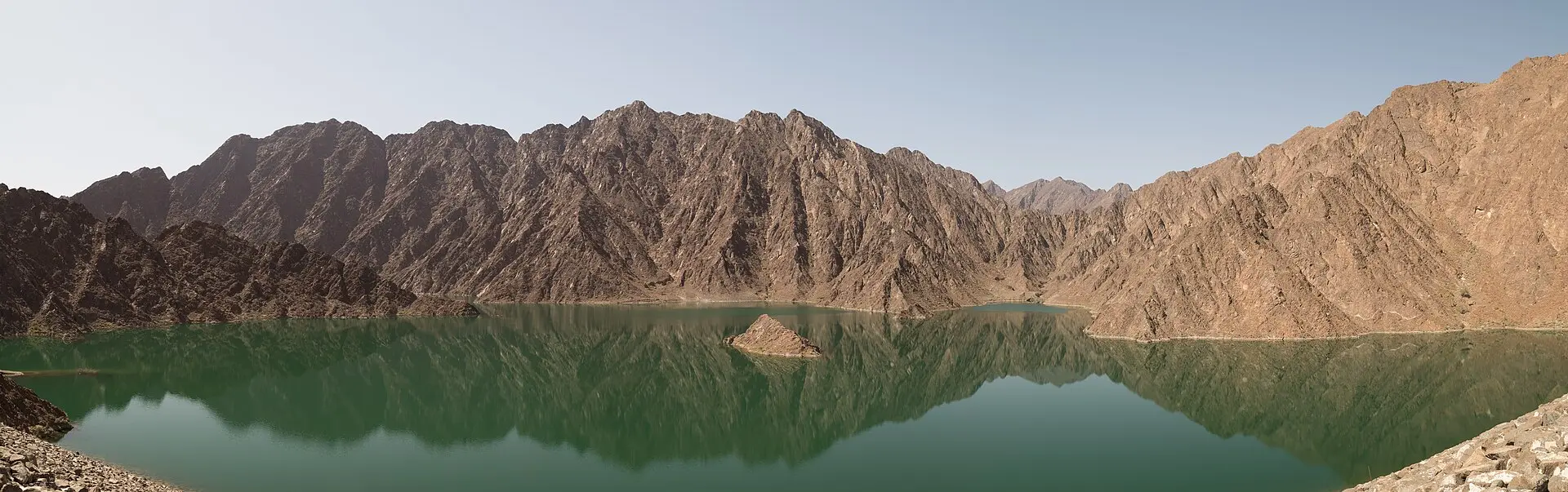 Hatta-Stausee mit glattem grünem Wasser, in dem sich schroffe braune Felsgrate des Hajar-Gebirges spiegeln, darüber klarer Himmel