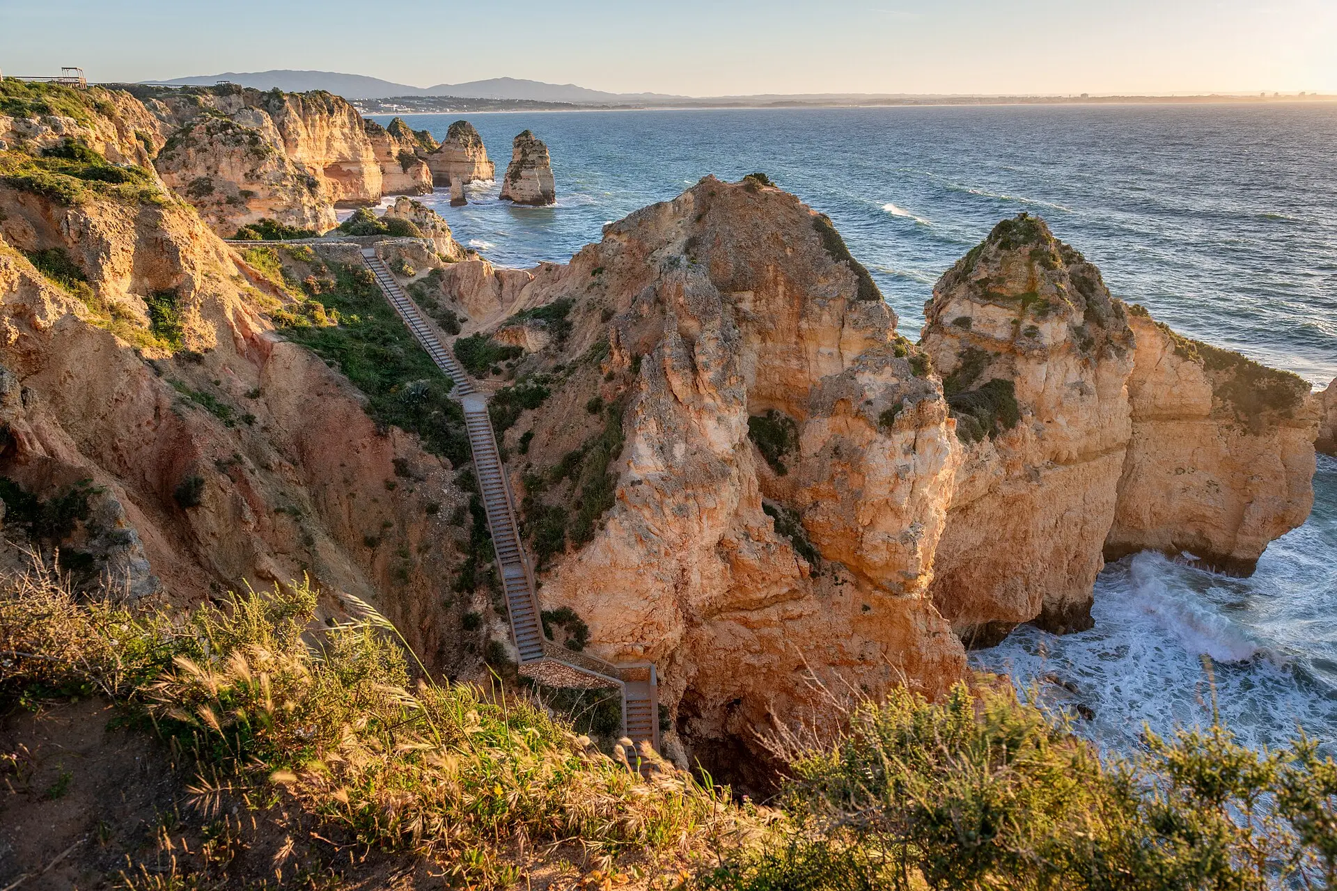 Lange Treppe mit Geländer zwischen ockerfarbenen Klippen an der Ponta da Piedade, Felsnadeln im Meer, Brandung, dahinter die Bucht von Lagos