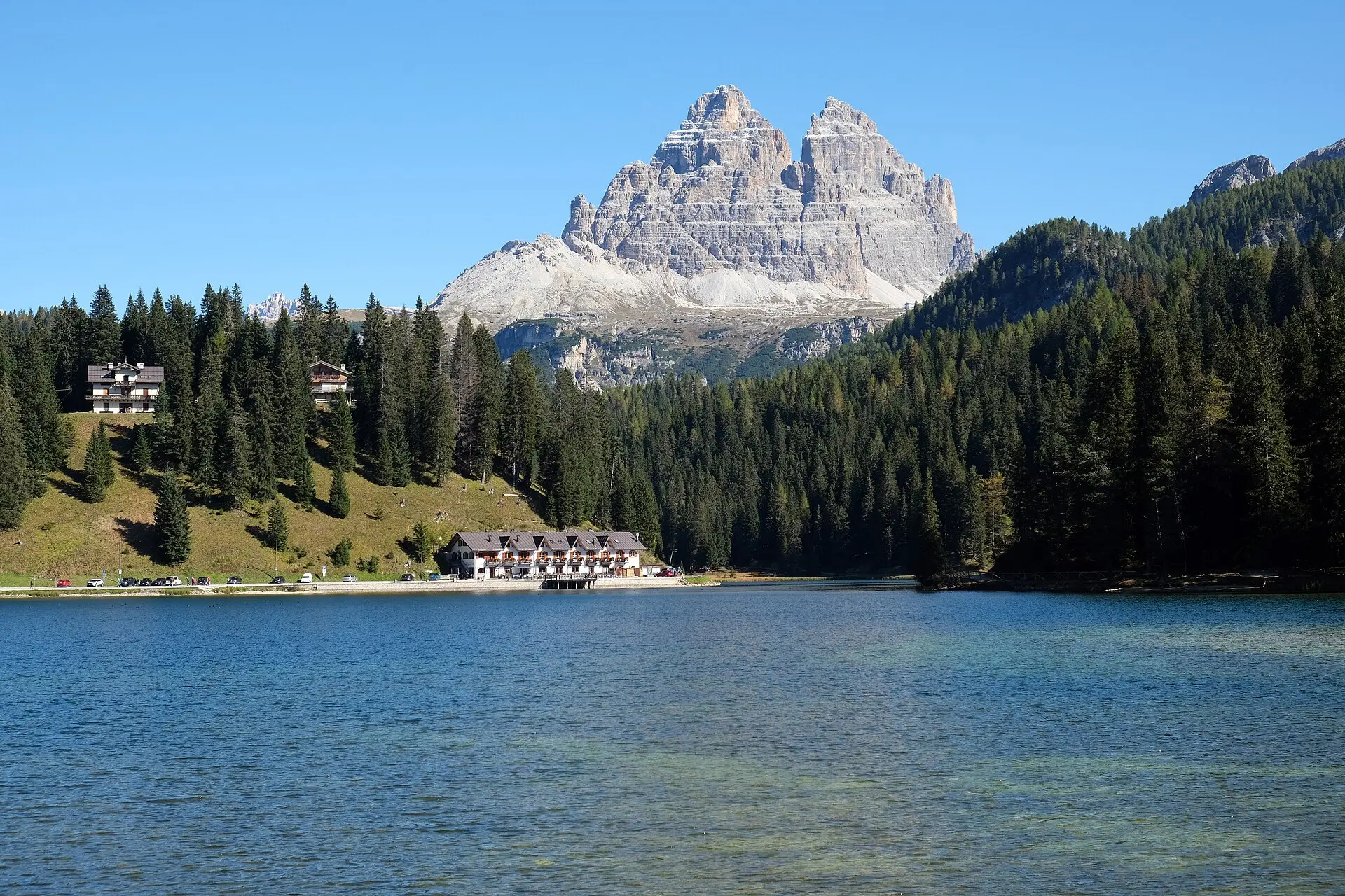 Misurinasee in den Dolomiten mit Nadelwald und einem Hotelbau am Ufer, dahinter die Felsgruppe der Drei Zinnen unter blauem Himmel