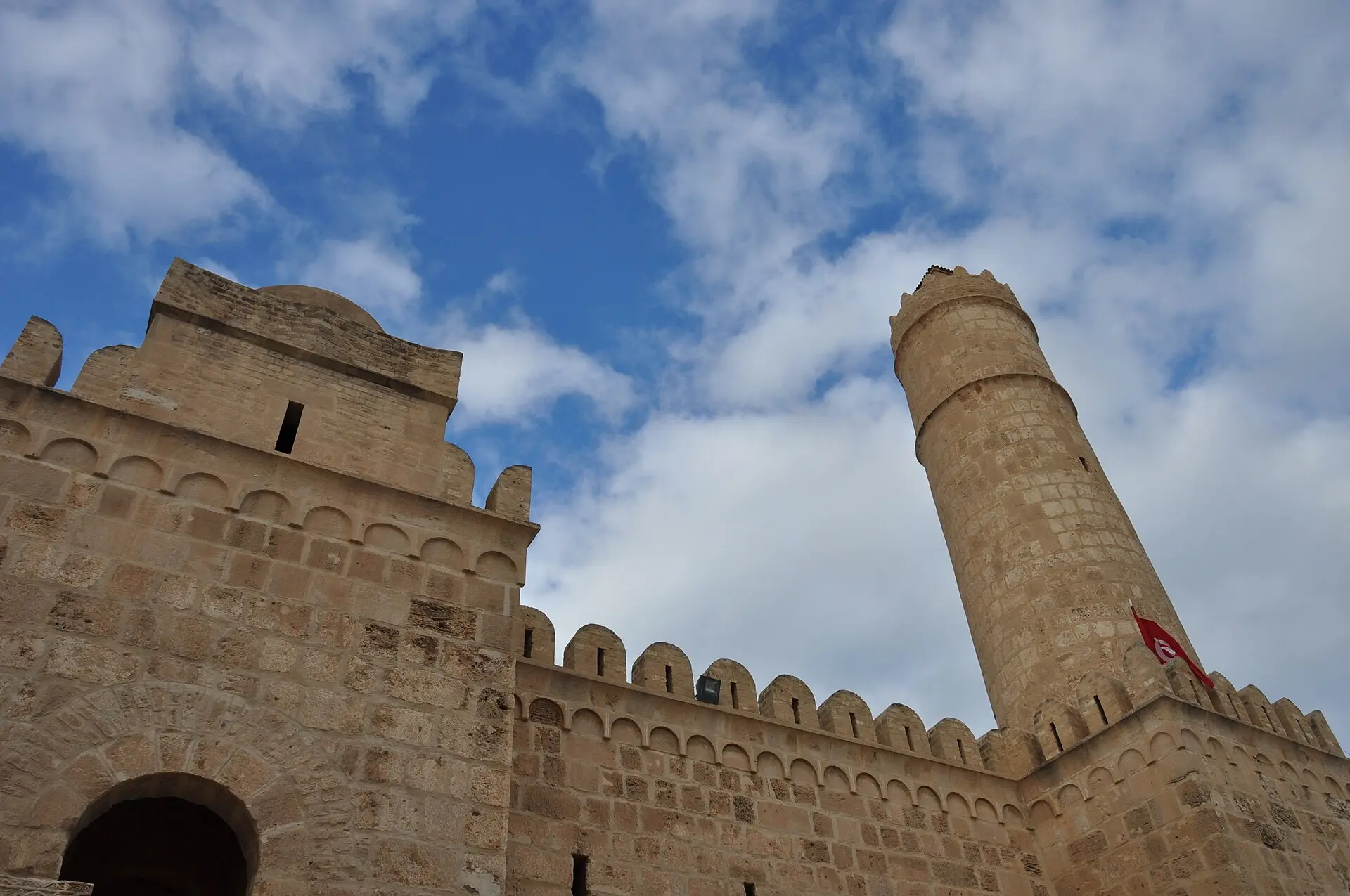 Ribat von Sousse von unten: Zinnenmauer mit Rundbogentor, runder Wachturm und rote tunesische Flagge vor blauem Himmel mit Wolken