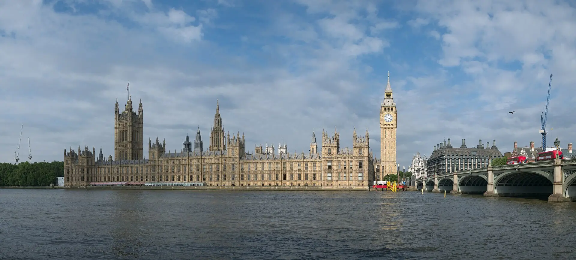 Palace of Westminster mit dem Elizabeth Tower und der Westminster Bridge, vom gegenüberliegenden Themseufer aus unter blauem Himmel mit Wolken
