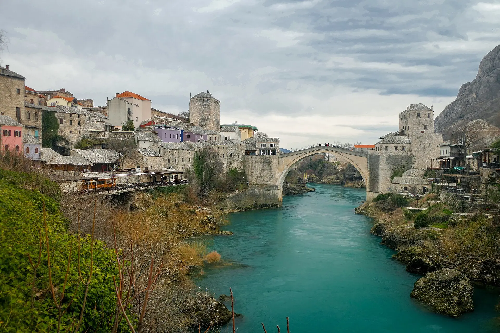 Alte Brücke von Mostar über der türkisfarbenen Neretva, links Steinhäuser der Altstadt, rechts ein steiler Felshang, bewölkter Himmel