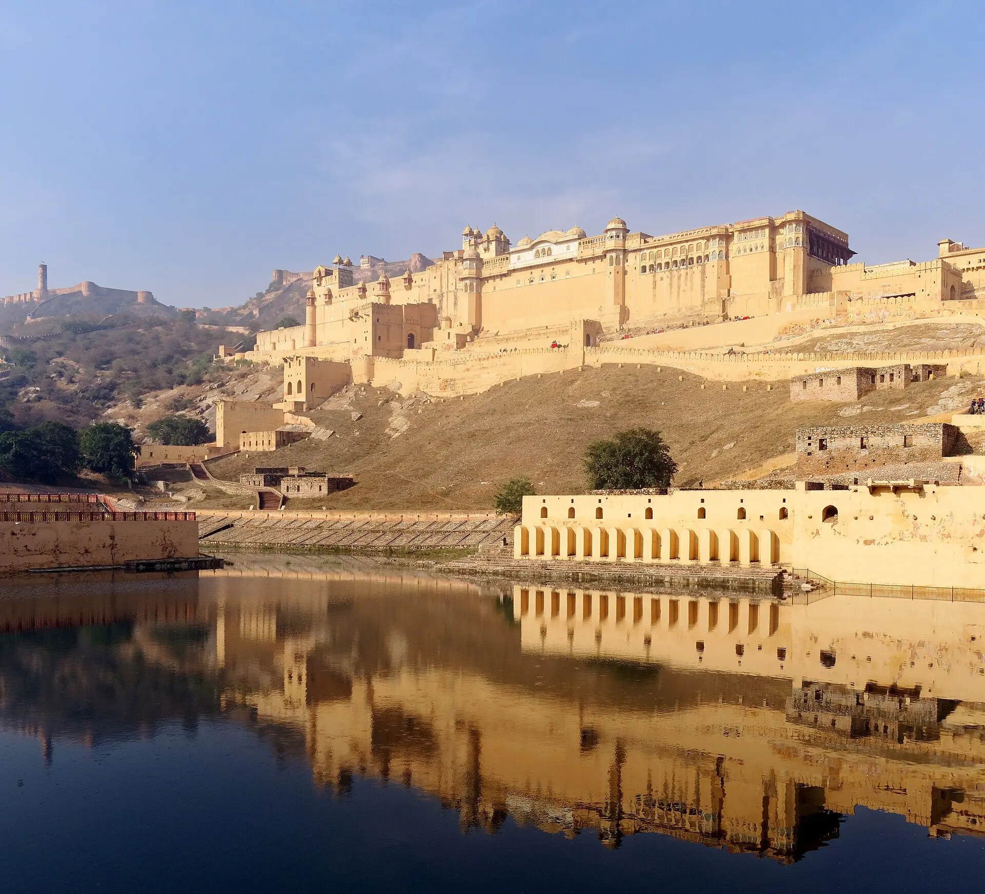 Ockergelbe Palastmauern des Amber Fort am Hang über dem Maota-See, die sich im Wasser spiegeln, oben links die Jaigarh-Festung