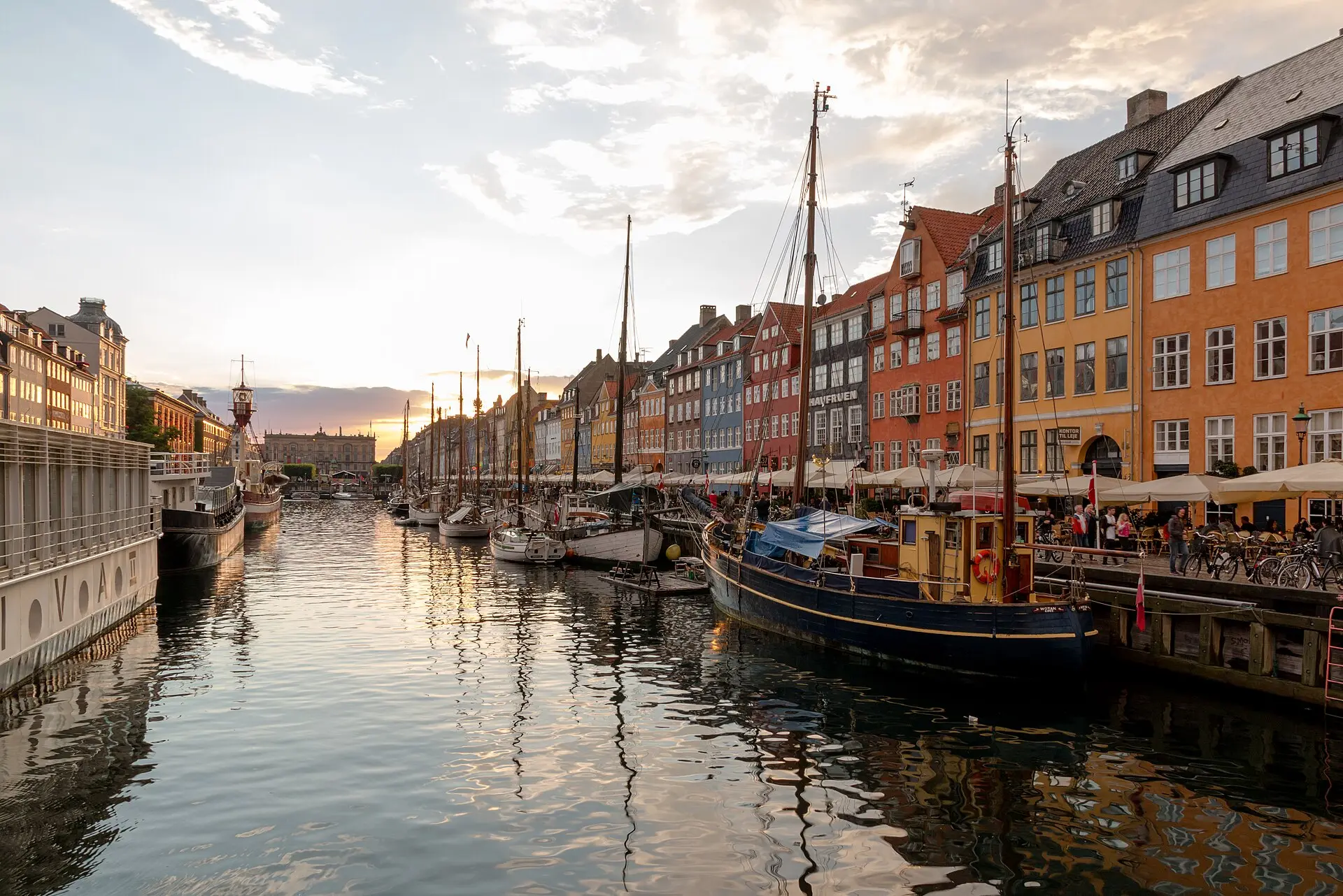 Nyhavn am Abend: bunte Giebelhäuser mit Café-Sonnenschirmen, im Kanal Holzsegler mit Masten, links ein weißes Schiff und ein Feuerschiff