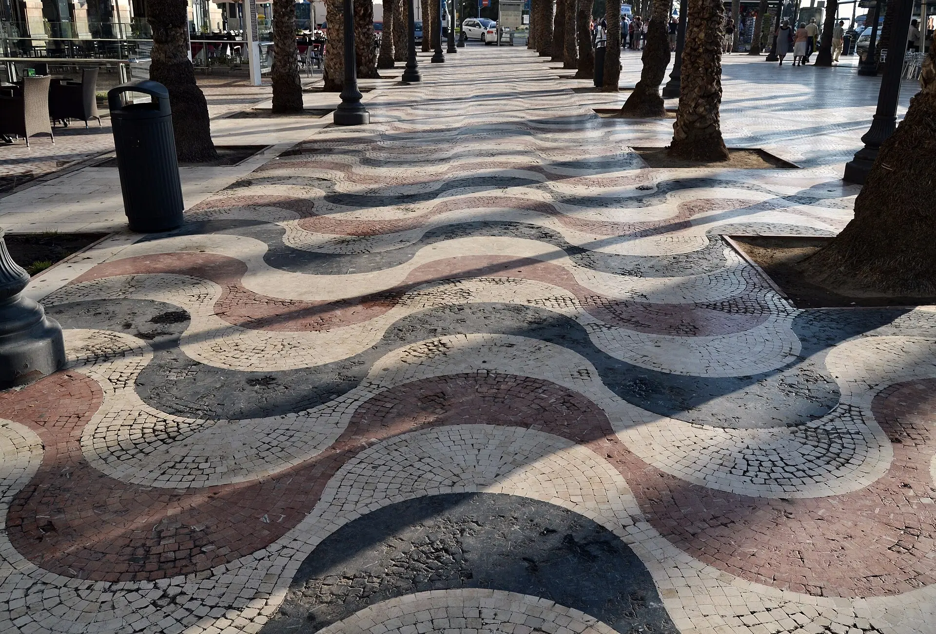 Wellenmosaik der Explanada de España aus kleinen Steinen in Rot, Anthrazit und Weiß, dazwischen Palmenstämme, links Café-Stühle