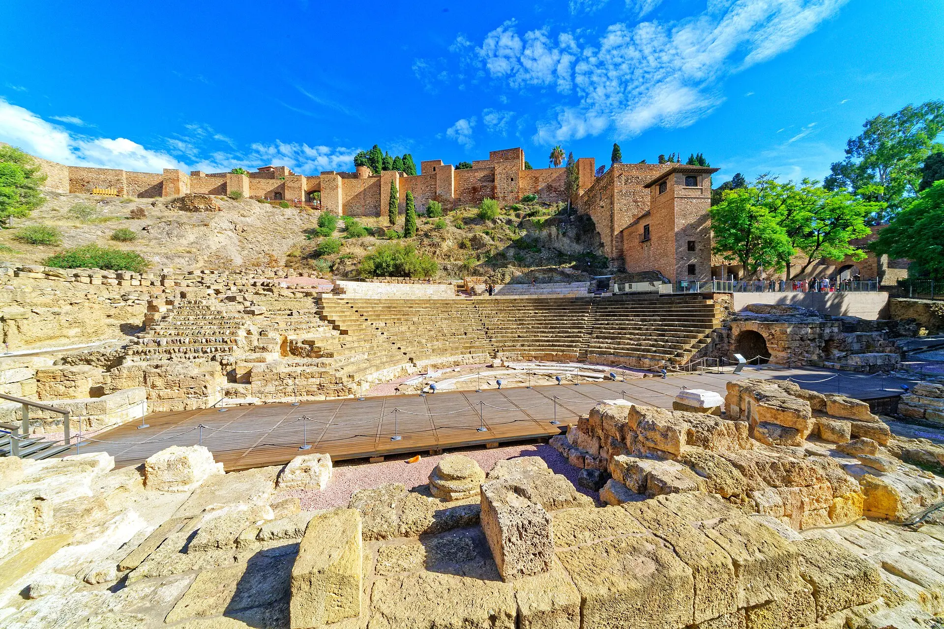Römisches Theater von Málaga mit Sitzstufen, Holzboden und Steinblöcken, darüber die Alcazaba mit Zypressen, rechts Besucher am Geländer