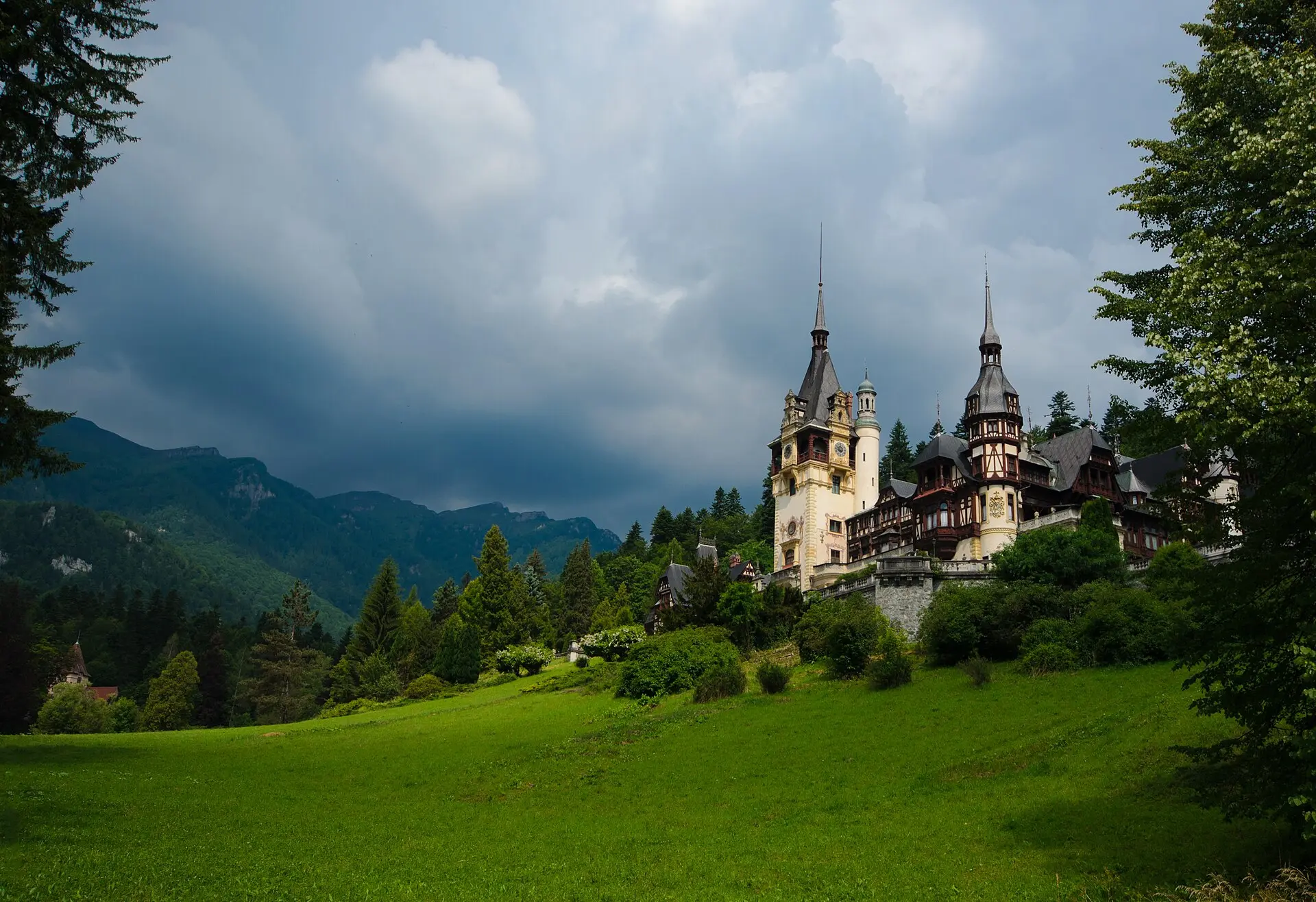 Schloss Peleș mit Türmen und Fachwerkgiebeln auf einem Hang über einer Wiese, links bewaldete Berghänge, dunkle Gewitterwolken