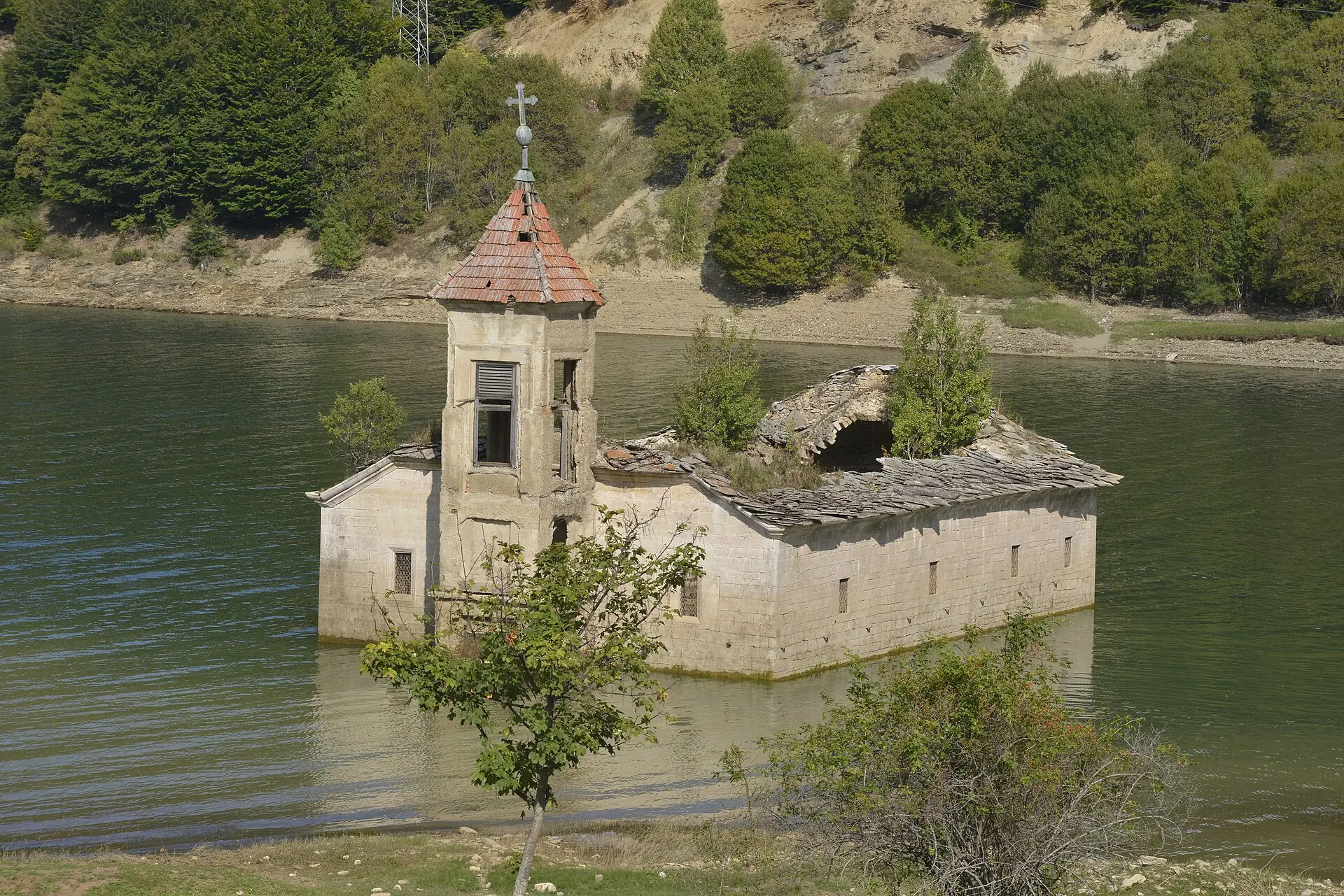 Kirche mit viereckigem Glockenturm und eingebrochenem Dach, die zum Teil im grünen Mavrovo-See steht, dahinter ein bewaldeter Hang