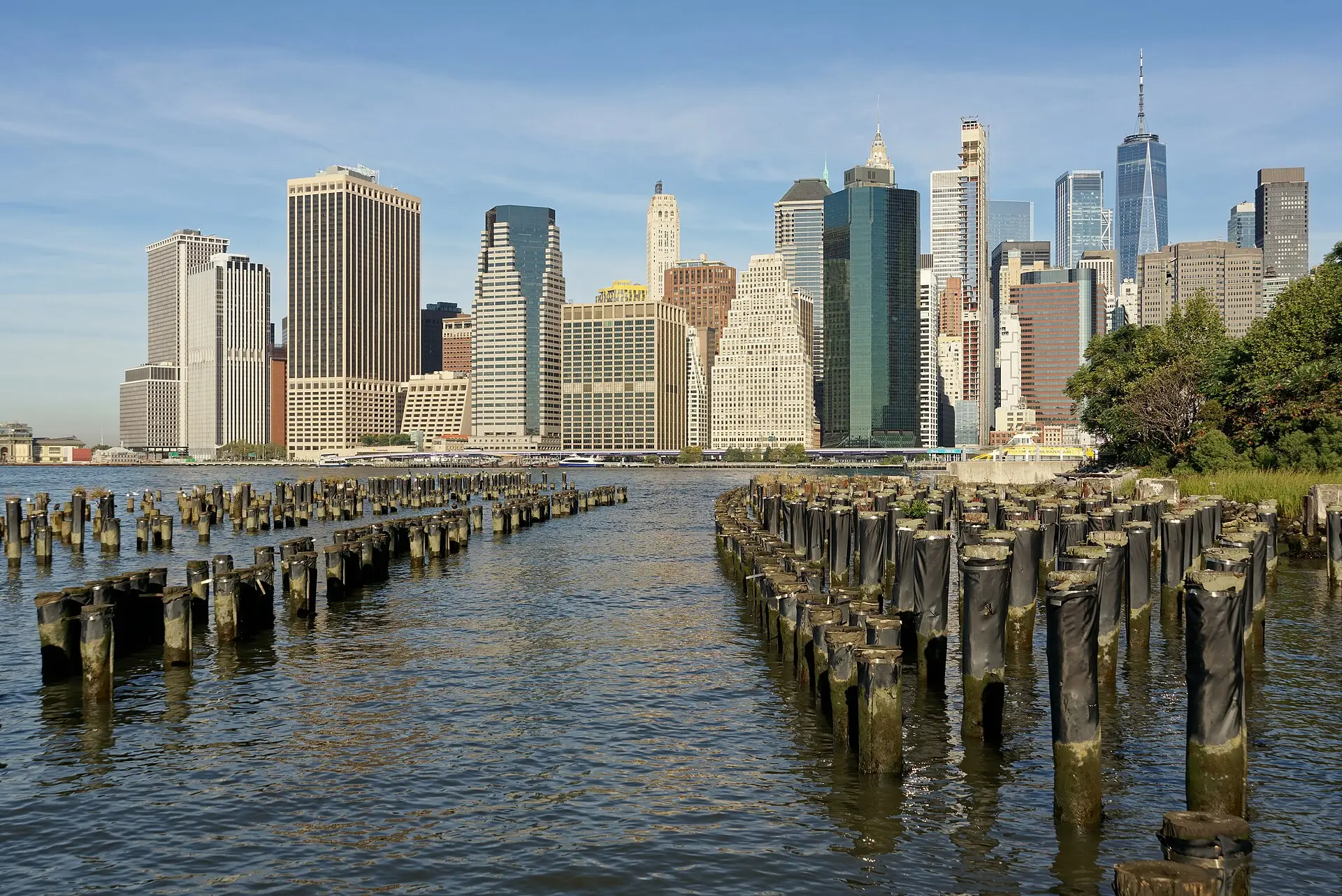 Hochhäuser des Financial District in Lower Manhattan von Brooklyn aus gesehen, davor alte Holzpfähle im Wasser und Bäume