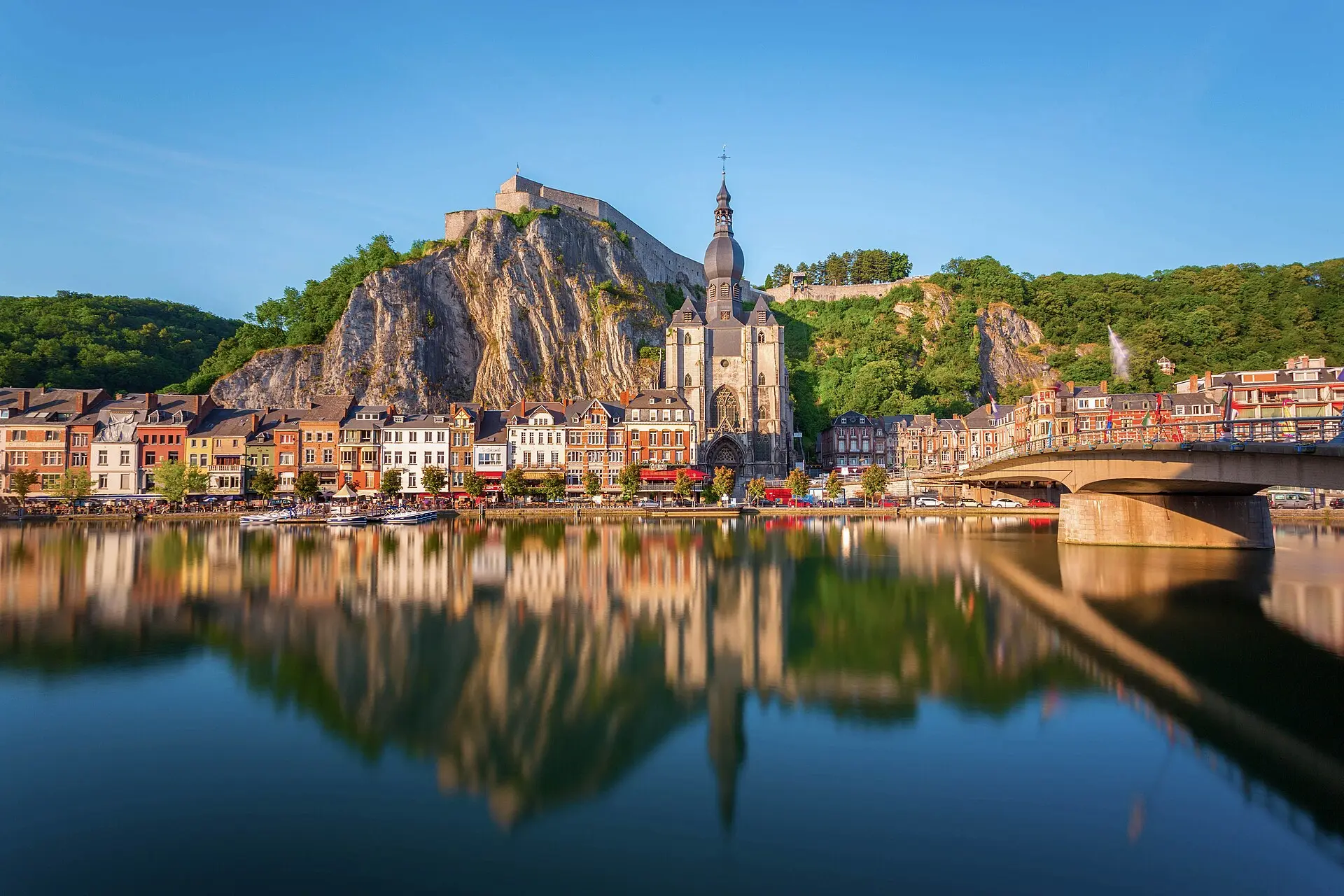 Dinant an der Maas: Stiftskirche mit zwiebelförmigem Turmhelm vor einer Felswand, oben die Zitadelle, die Stadt spiegelt sich im Fluss