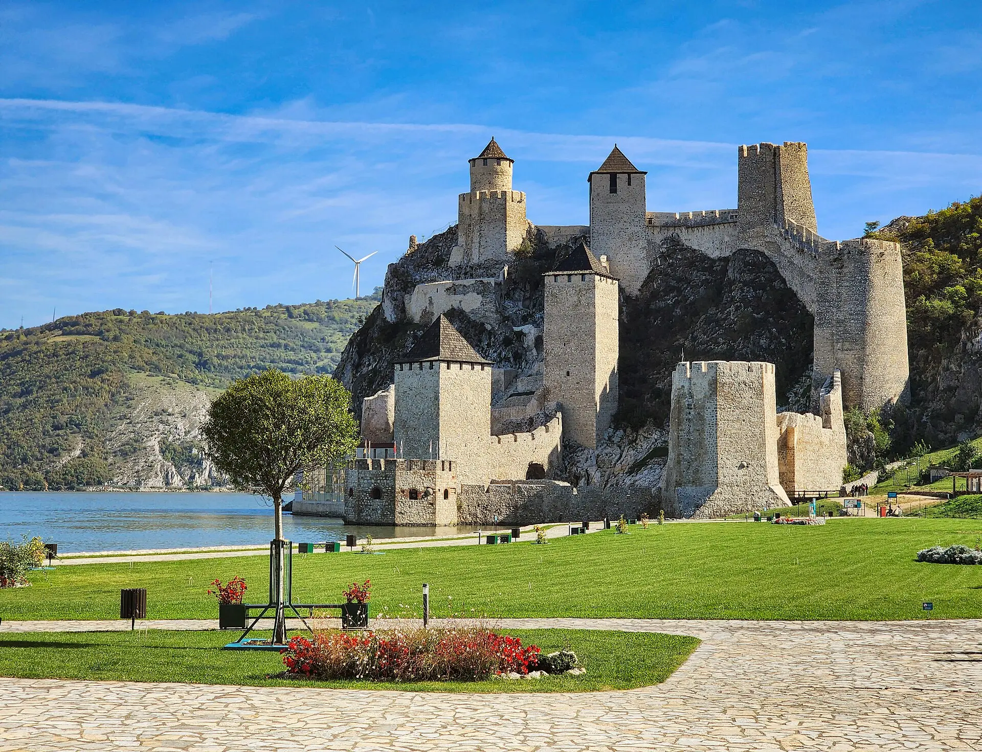 Festung Golubac an der Donau: Steintürme mit spitzen Dächern am Felshang, davor Rasen, ein gepflasterter Weg und ein Blumenbeet