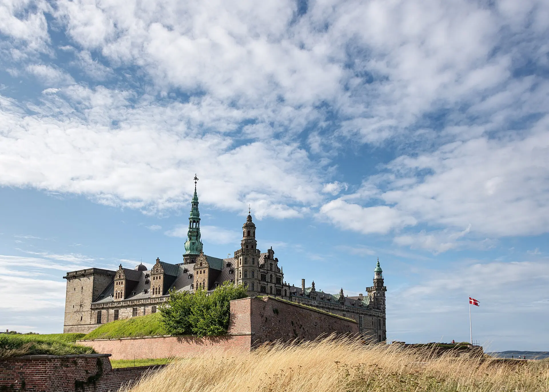 Schloss Kronborg in Helsingør: Sandsteinfassade mit grünem Kupferturm hinter einer Backsteinmauer, davor hohes trockenes Gras