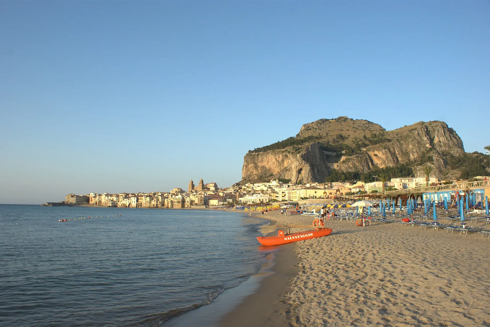 Sandstrand von Cefalù im Abendlicht mit orangem Rettungsboot, blauen Schirmen und Badegästen, dahinter Altstadt, Domtürme und Rocca