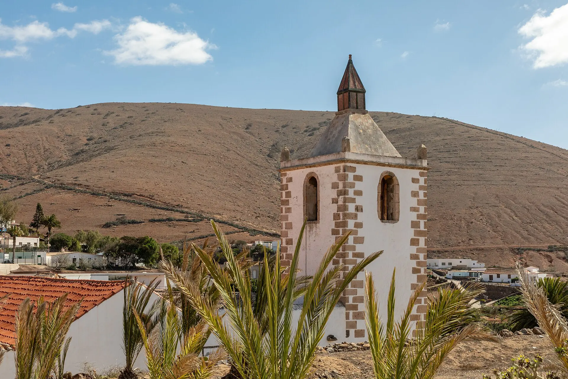 Weiß verputzter Glockenturm der Kirche Santa María in Betancuria, davor Palmwedel und ein rotes Ziegeldach, dahinter kahle braune Hügel