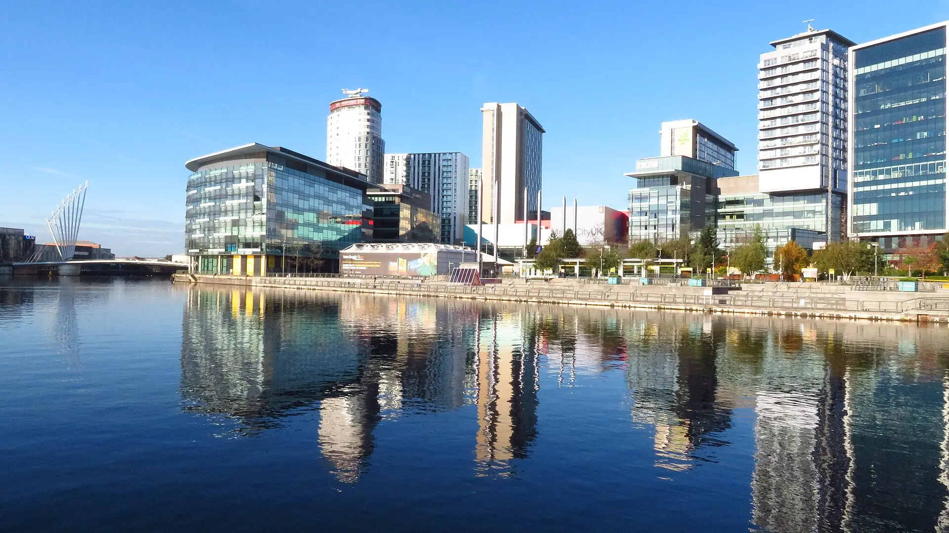 Glasfassaden und Hochhäuser von MediaCityUK spiegeln sich im Hafenbecken von Salford Quays, links eine weiße Fußgängerbrücke
