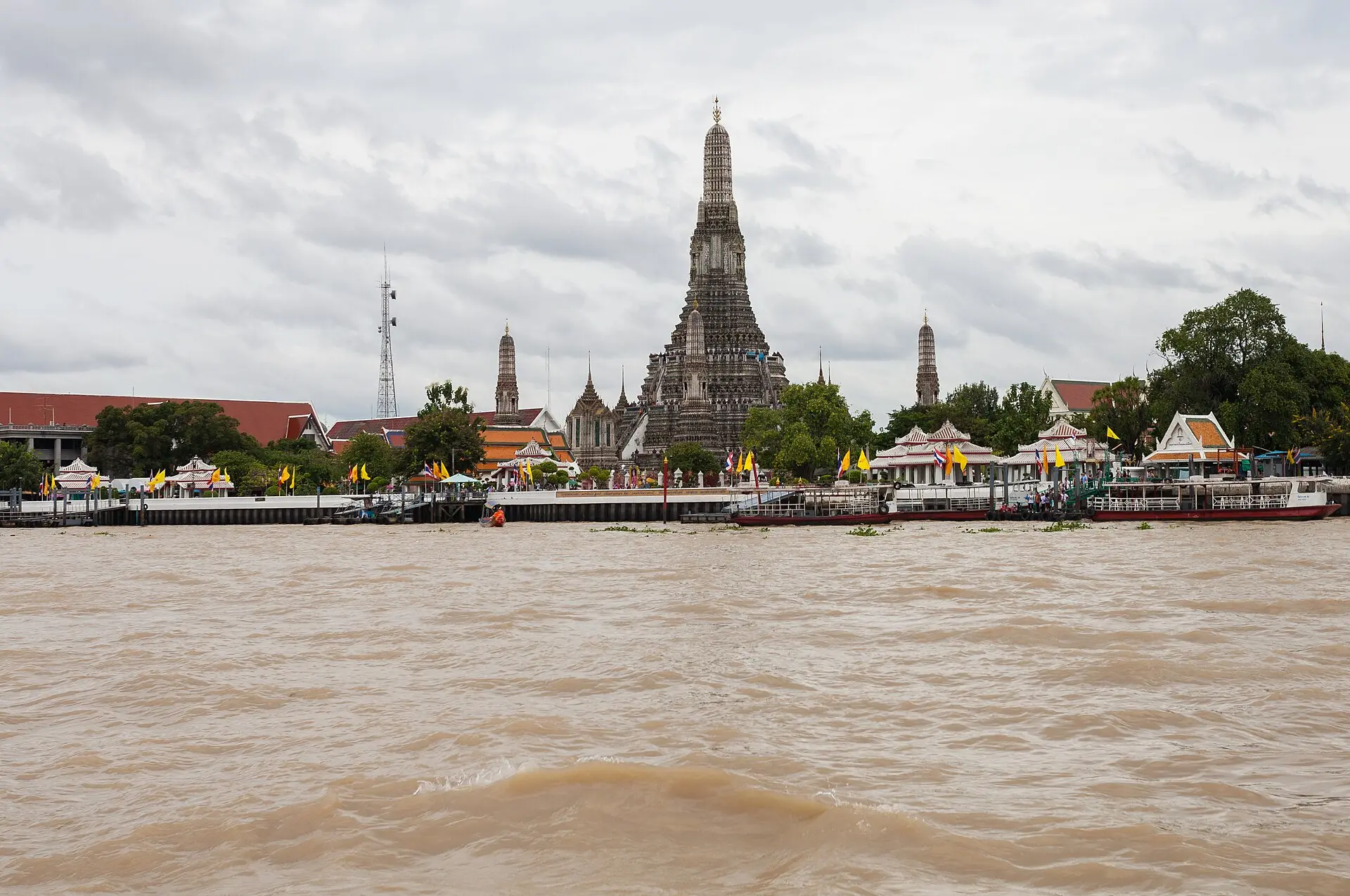 Wat Arun vom Chao Phraya aus: hoher Hauptprang mit zwei kleineren Prangs, davor weiße Pavillons, ein Anleger und Boote