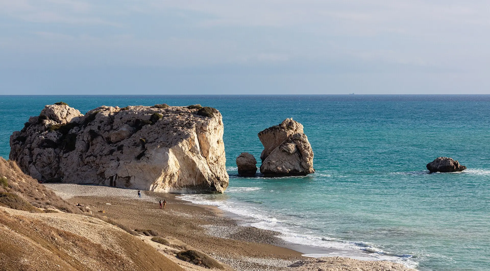 Petra tou Romiou von einem Hang aus: großer heller Felsen am Ufer, ein Felsturm im türkisblauen Meer, davor Strand aus Sand und Kies