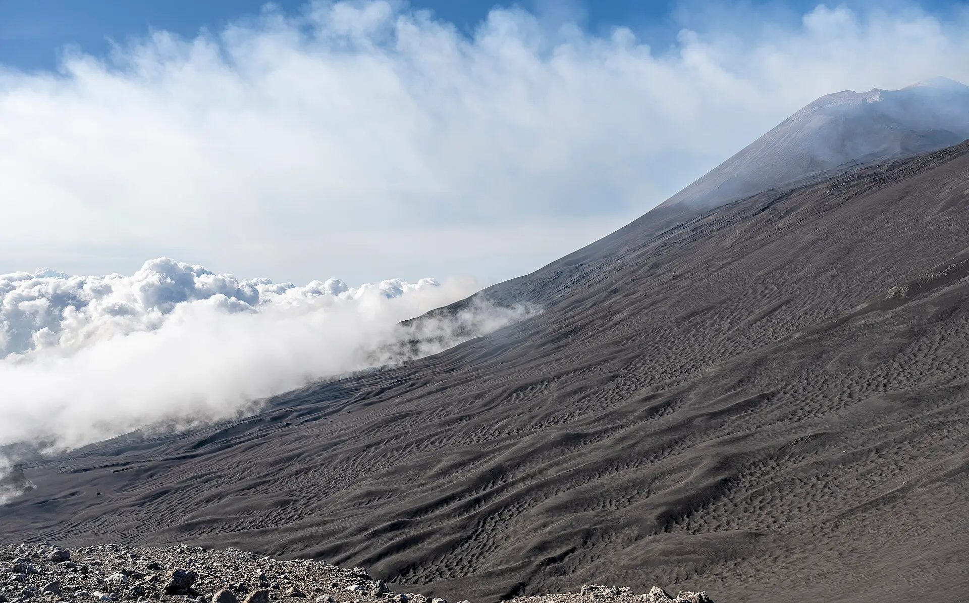 Flanke des Ätna auf rund 2.800 m: schwarze, gewellte Vulkanasche, rechts oben ein Gipfelkegel, links ein Wolkenmeer