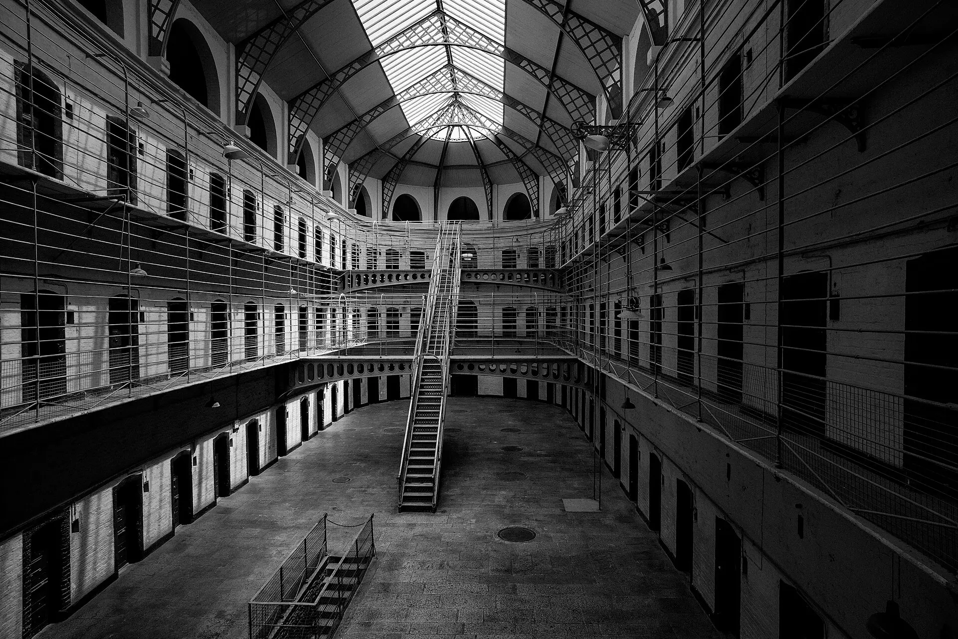Schwarzweißbild des leeren Ostflügels von Kilmainham Gaol: drei Etagen Zellentüren an Galerien, Metalltreppe und Glasdach