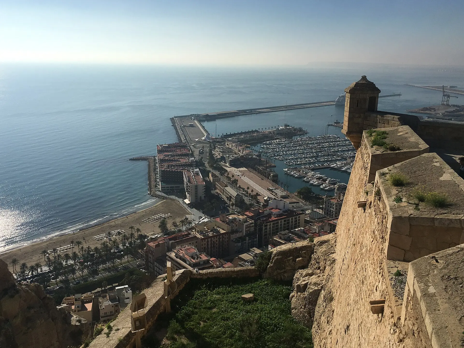 Blick von der Burgmauer des Castillo de Santa Bárbara auf Postiguet-Strand, Palmen, Jachthafen und Hafen von Alicante, rechts ein Wachturm