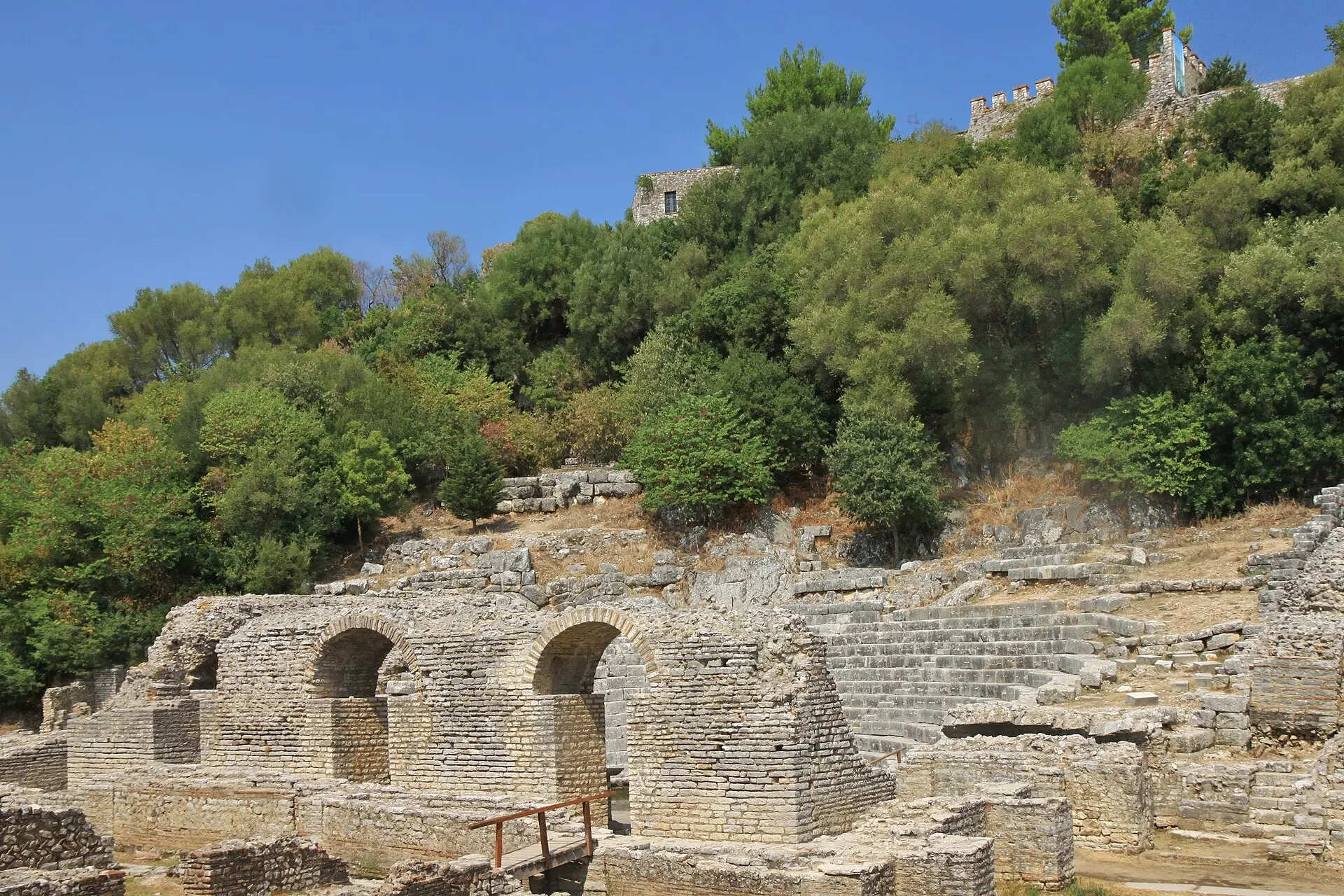 Antikes Theater von Butrint mit steinernen Sitzreihen und Mauerbögen, darüber ein bewaldeter Hügel mit Zinnenmauer