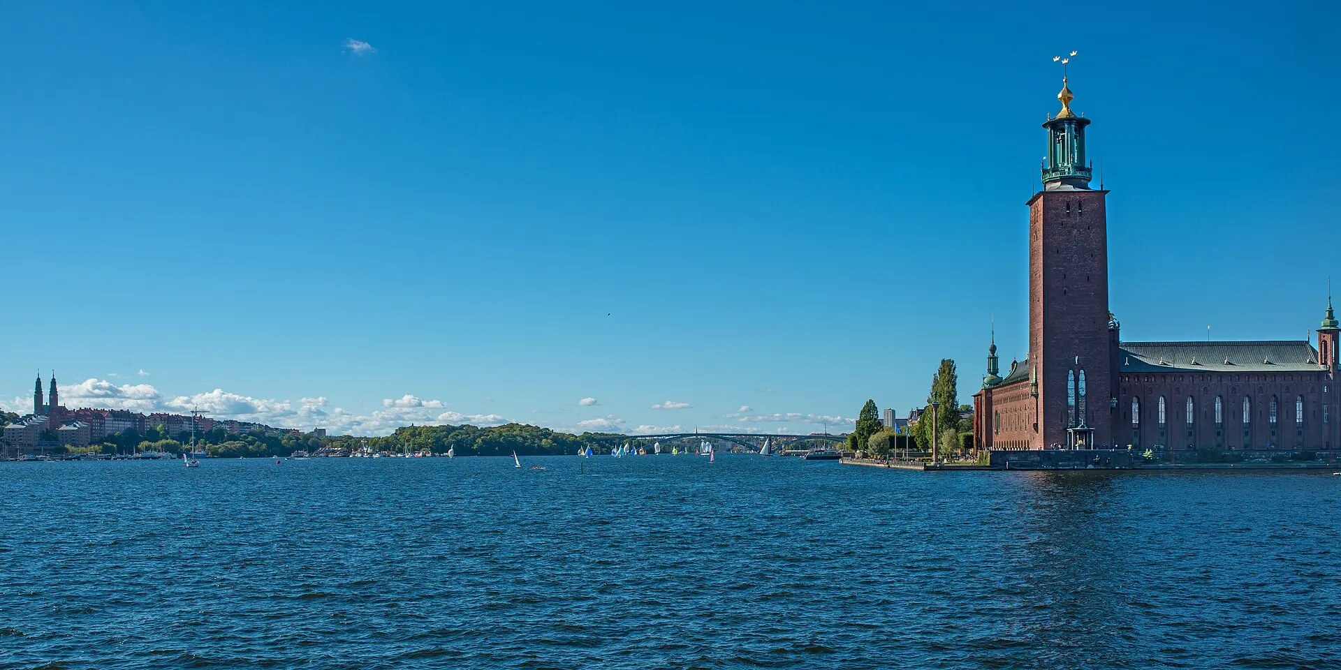 Das Rathaus mit Turm und drei goldenen Kronen am Wasser, links Segelboote auf dem Riddarfjärden, dahinter die Västerbron