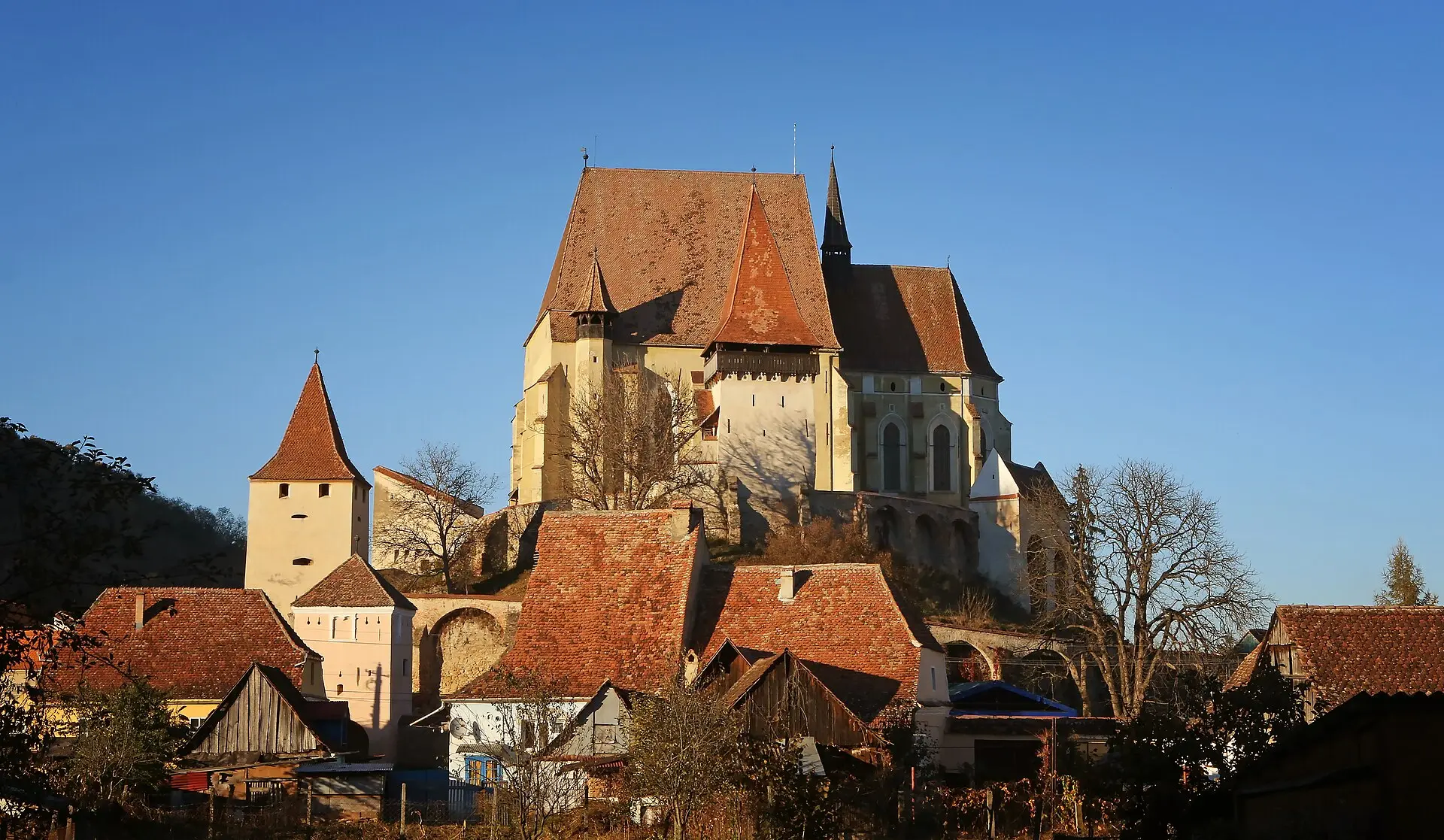 Kirchenburg Biertan auf einem Hügel in tief stehender Sonne: Kirche mit steilem Ziegeldach, Wehrtürme und Mauerring, davor Dächer des Dorfes