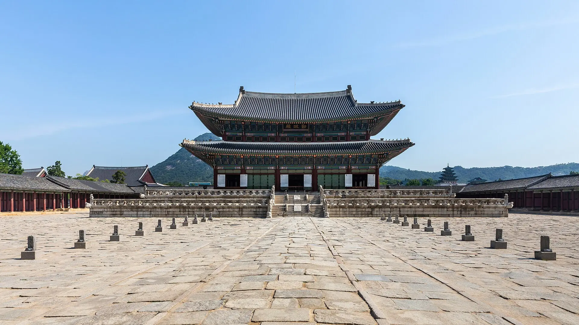 Thronhalle Geunjeongjeon im Gyeongbokgung in Seoul: Holzhalle mit geschwungenen Dächern auf Steinterrasse, davor leerer Hof, hinten Berge