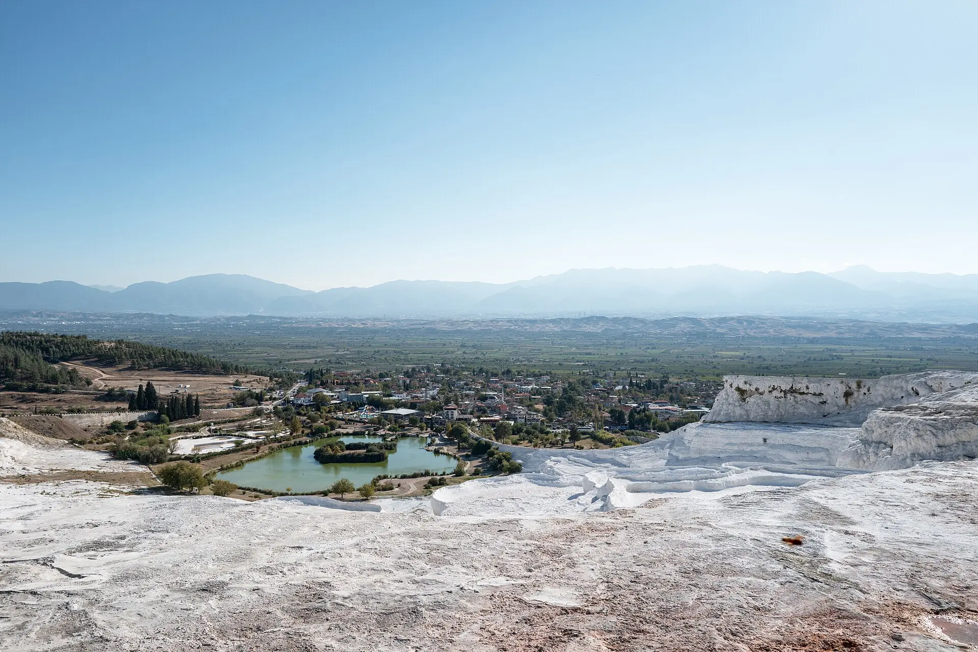 Weiße Kalksinterterrassen von Pamukkale, darunter ein grüner Teich und das Dorf, dahinter eine weite Ebene und eine blaue Bergkette