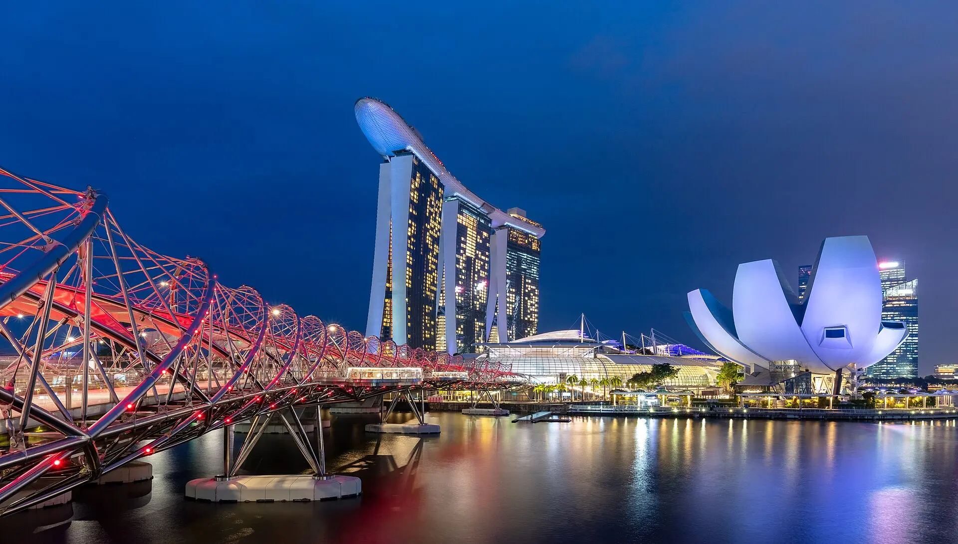 Marina Bay zur blauen Stunde: rot beleuchtete Helix-Brücke, die drei Türme des Marina Bay Sands und das weiße ArtScience Museum
