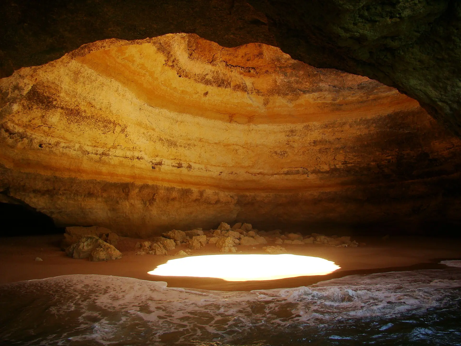 Innenraum der Benagil-Grotte mit gewölbter Decke aus gelb-orangen Kalkschichten, Sandstrand, Lichtfleck und Wellen
