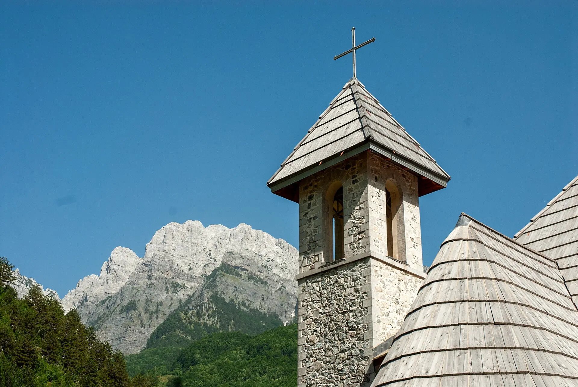 Steinerner Glockenturm der Kirche von Theth mit Schindeldach, dahinter schroffe Kalkgipfel und Nadelwald