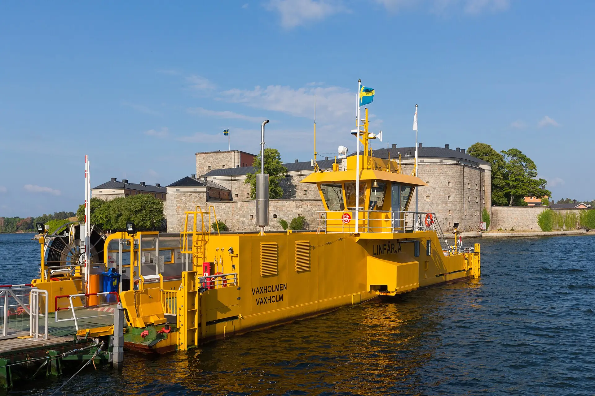 Gelbe Seilfähre mit schwedischer Flagge am Anleger in Vaxholm, dahinter die graue Steinfestung Vaxholm unter blauem Himmel