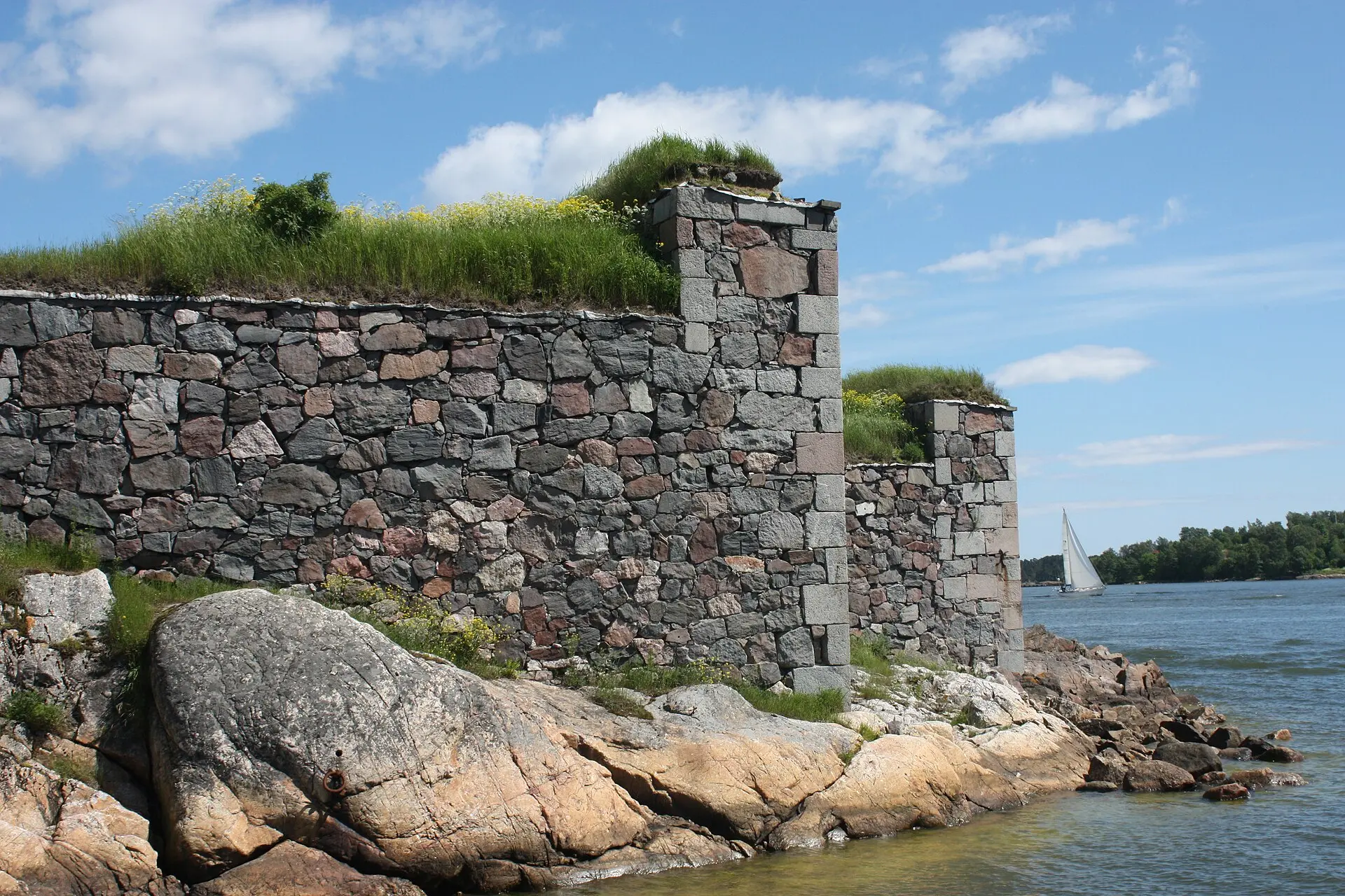 Graue Bruchsteinmauern der Festung Suomenlinna mit Gras obenauf, davor rosa Granitfelsen, rechts Meer und ein Segelboot
