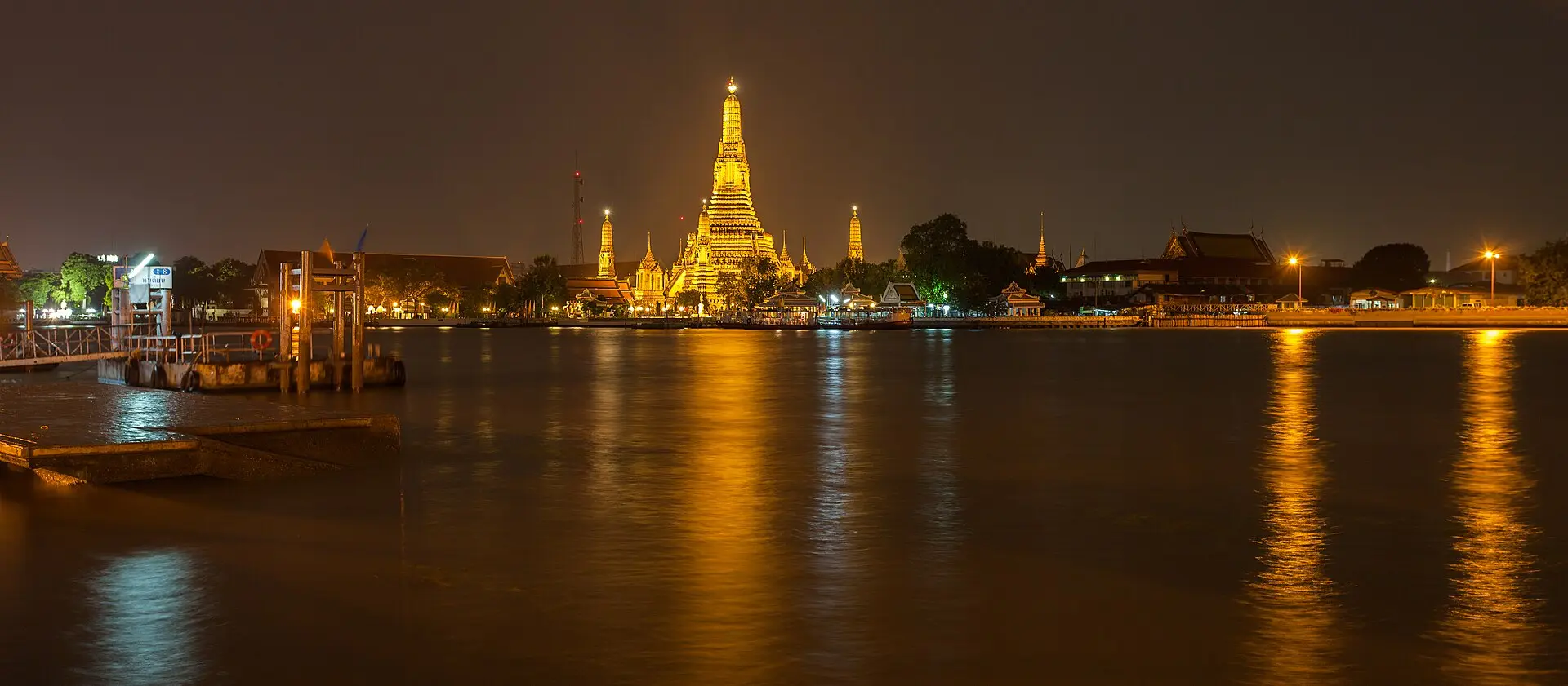 Angestrahlter Hauptprang des Wat Arun bei Nacht, goldgelb gespiegelt im Chao Phraya, links ein Bootsanleger mit Geländer