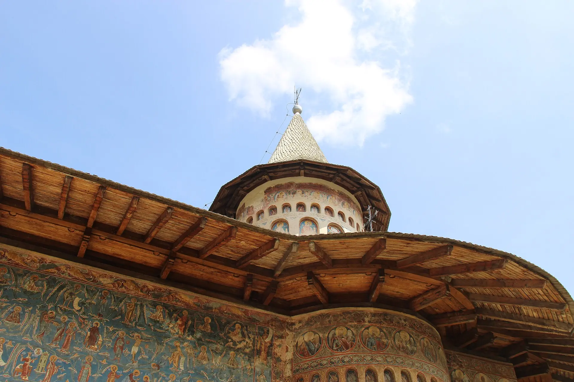 Kloster Voroneț von unten: bemalte Außenwand mit Heiligenfiguren auf blauem Grund, breites Holzdach, Turm mit Schindelhelm