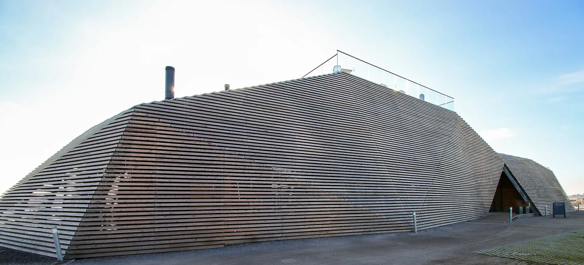 Außenansicht der Sauna Löyly: Gebäude aus schräg gestellten Holzlatten mit Metallschornstein und Glasgeländer auf dem Dach