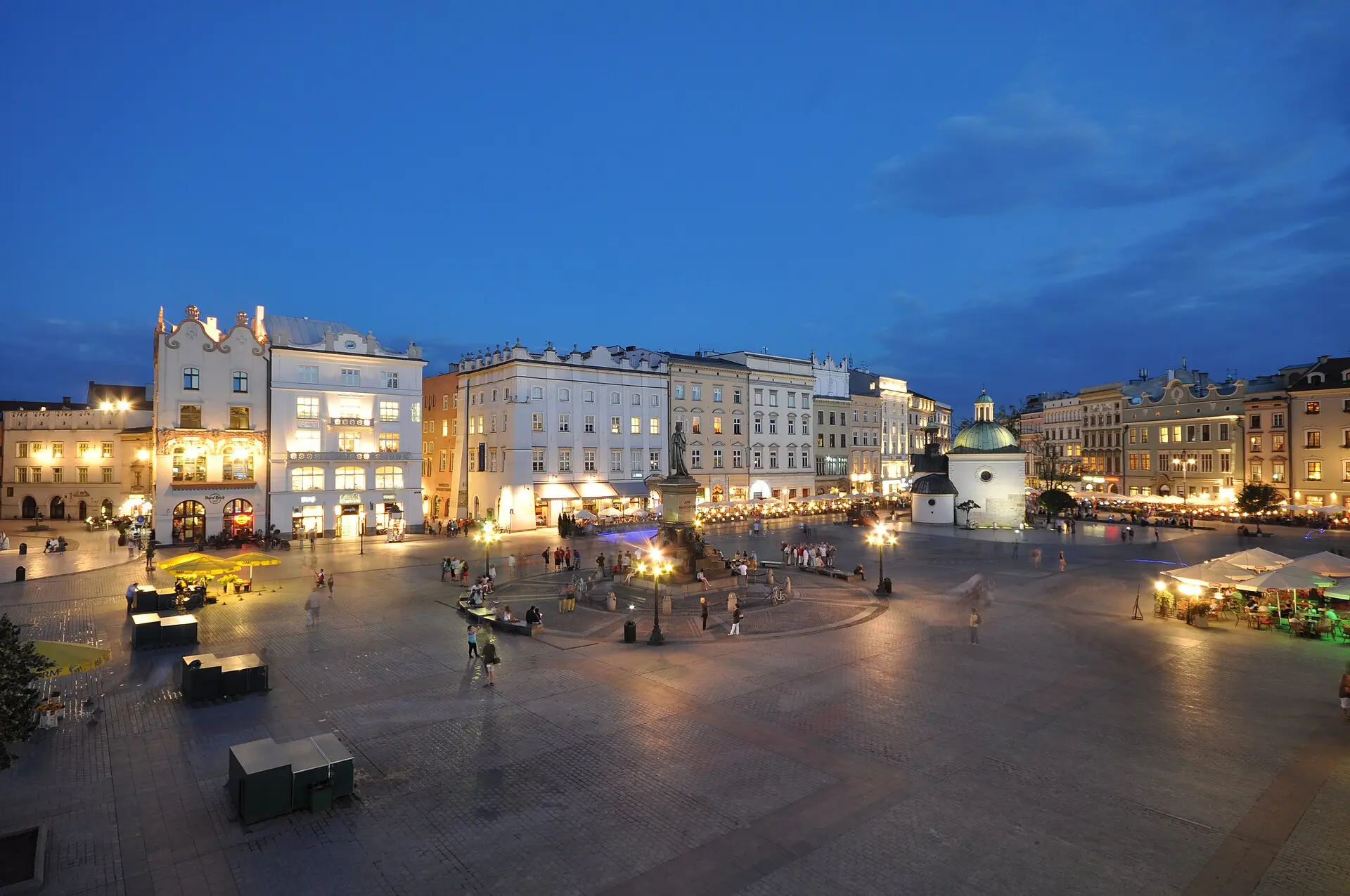 Hauptmarkt in Krakau zur blauen Stunde mit beleuchteten Bürgerhäusern, Denkmal in der Platzmitte und kleiner Kirche mit grüner Kuppel