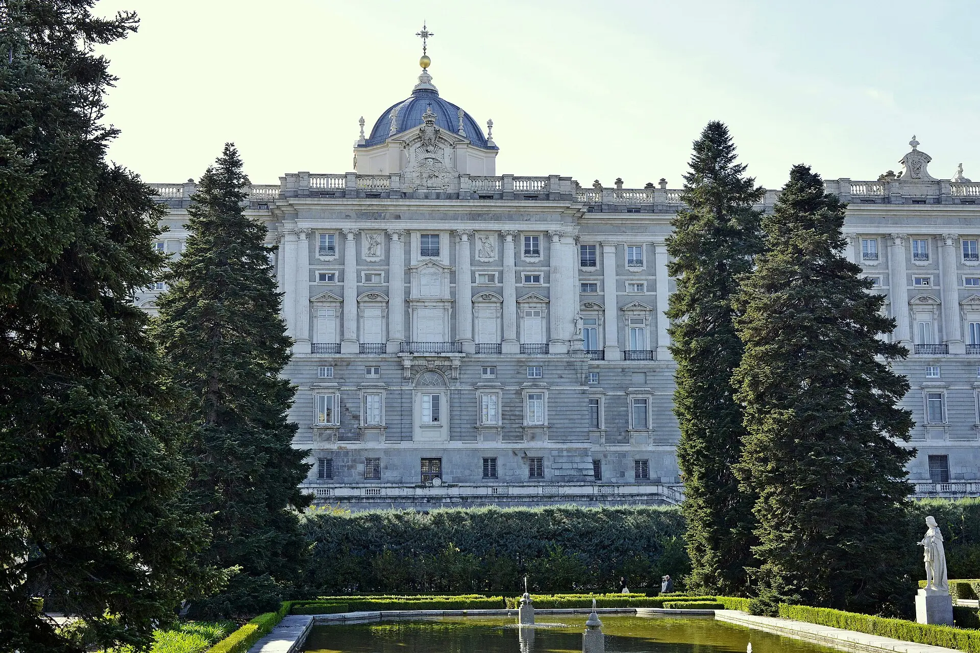 Nordfassade des Palacio Real in Madrid mit Kuppel, davor die Sabatini-Gärten mit hohen Nadelbäumen, Hecken und einem Wasserbecken