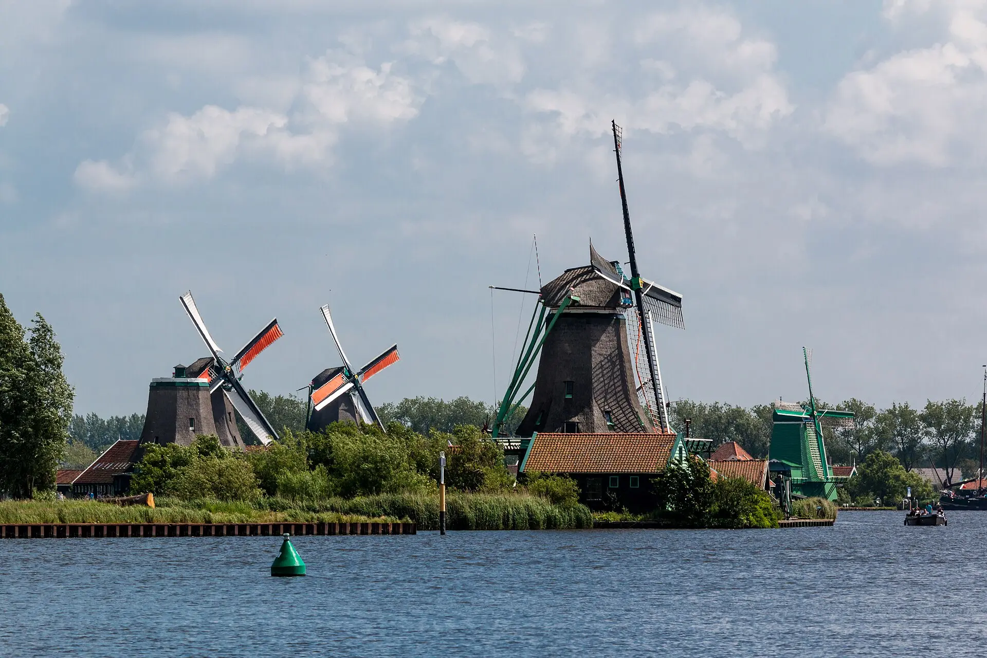 Fünf Windmühlen der Zaanse Schans am Ufer der Zaan, eine davon ohne Flügel, vom Wasser aus gesehen, mit Schilf und grüner Boje