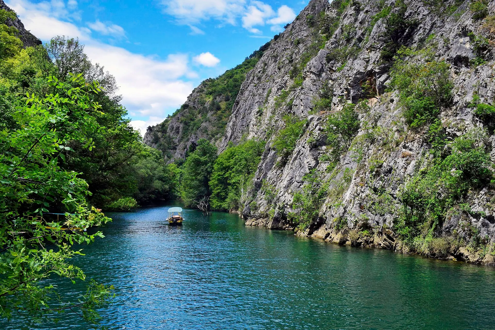 Steile graue Felswand über grün-blauem Wasser in der Matka-Schlucht, in der Mitte ein kleines Ausflugsboot mit weißem Dach
